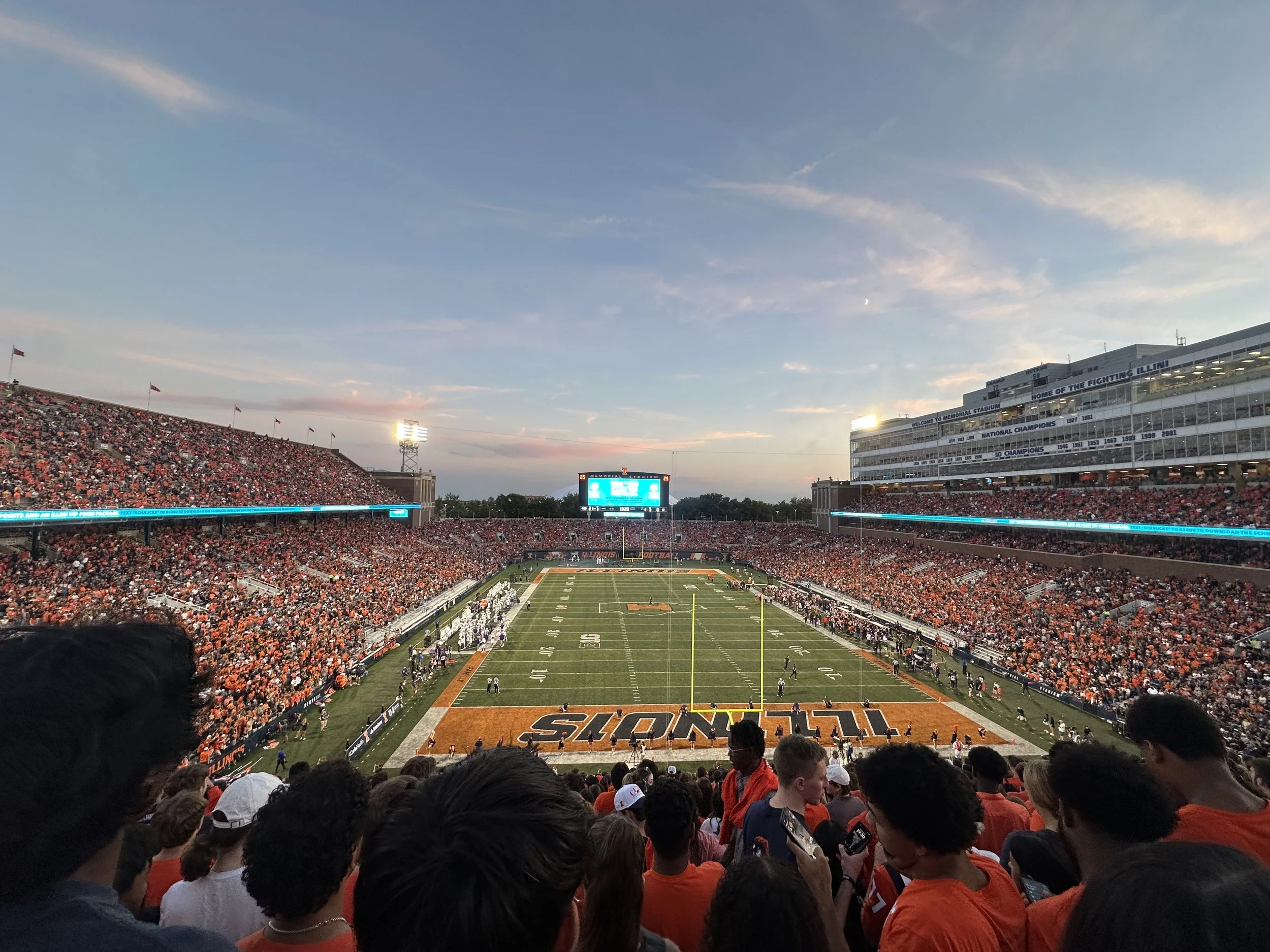 View of a football stadium filled with fans, with players on the field before a game at sunset.