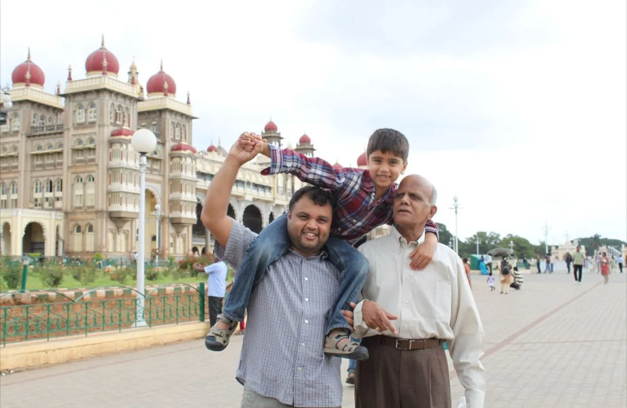 Three males, a child sitting on an adult's shoulders, smiling and playing, in front of a historic building with domes, in a public square with people walking around.
