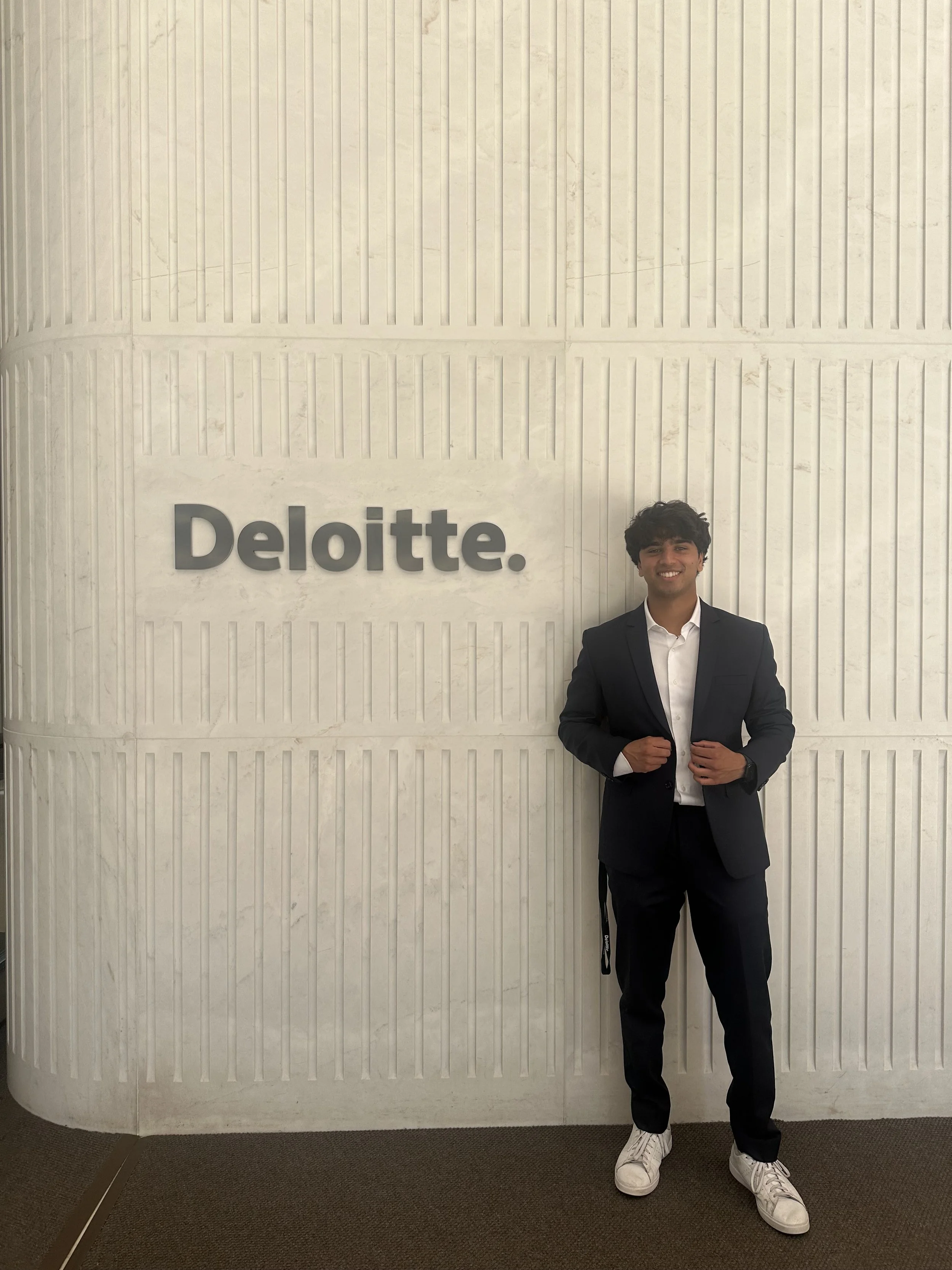 A young man in a black suit and white sneakers standing in front of a white textured wall with a 'Deloitte.' sign, smiling and adjusting his suit jacket.