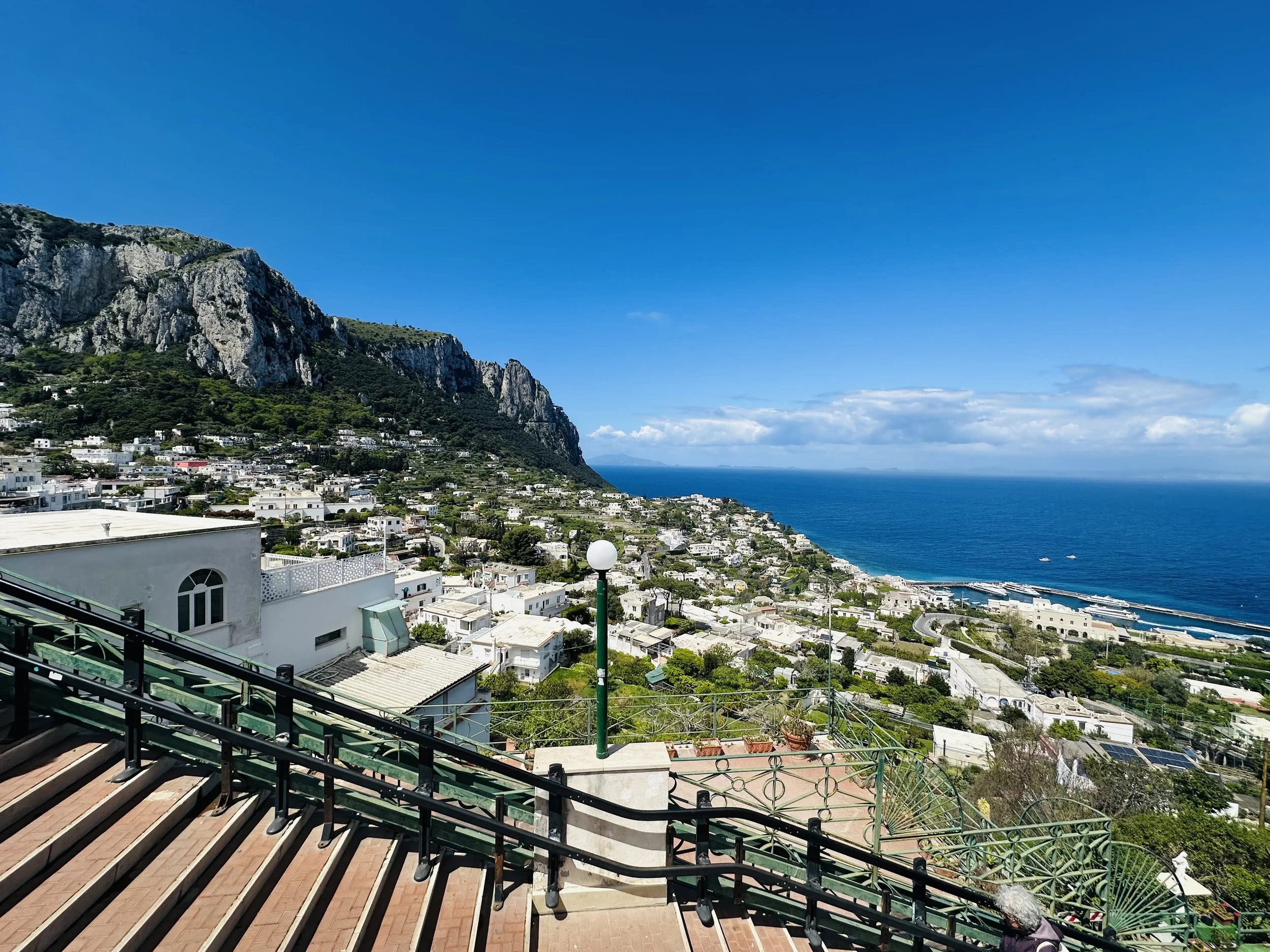 Scenic view of a coastal town with hillside white buildings, rocky mountain, and blue ocean under a partly cloudy sky, taken from a balcony with railing and outdoor seating.