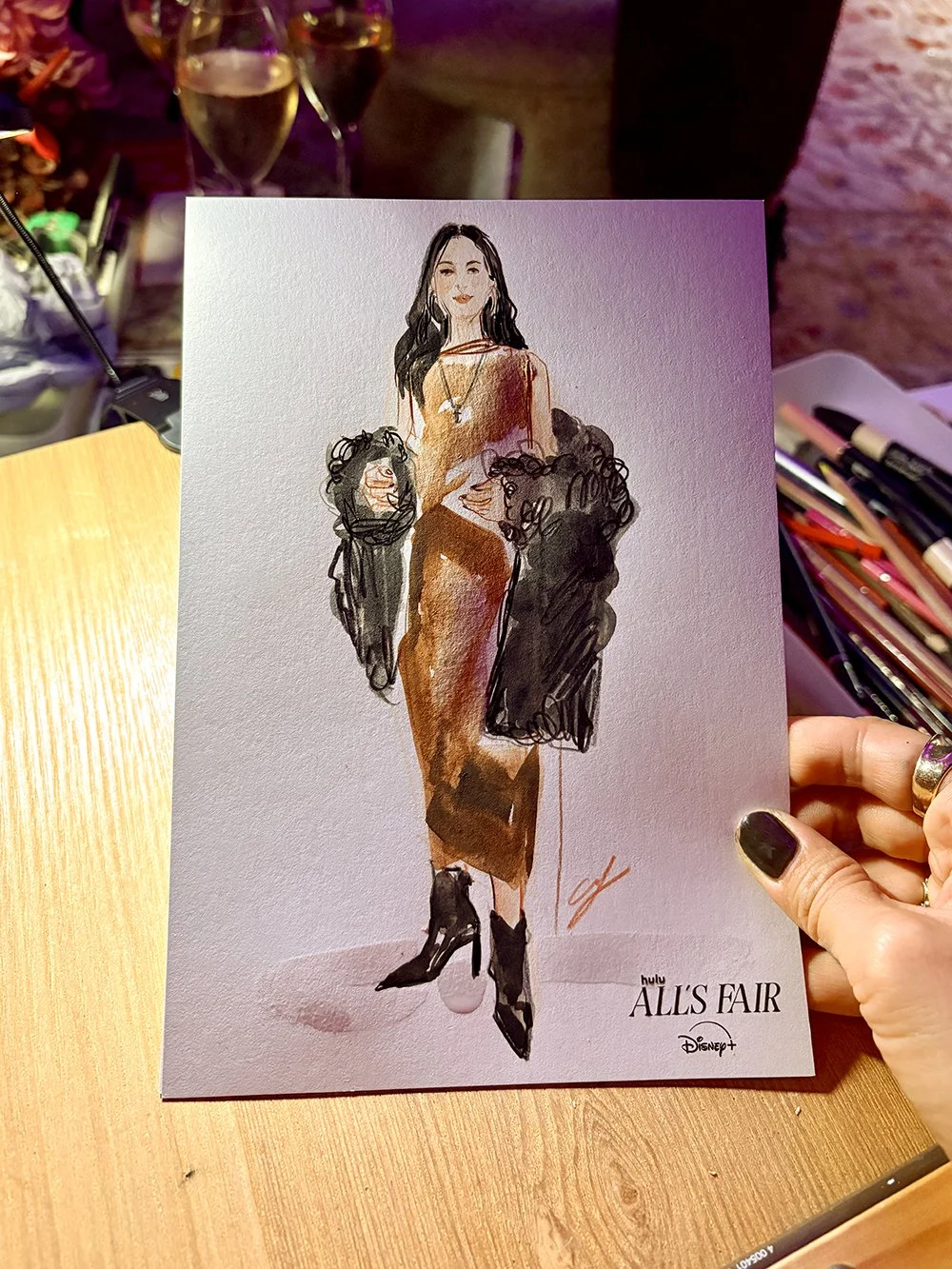 A hand holding a promotional card for Hulu's show 'All's Fair' featuring an illustration of a woman in a brown dress and black heels, flanked by two women with curly hair, in a setting with makeup and art supplies nearby.