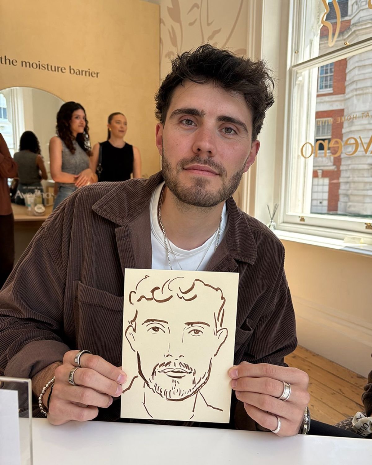 A man with dark hair and a beard holding a drawing of a man's face, sitting indoors near a window with a city view, with several people in the background.