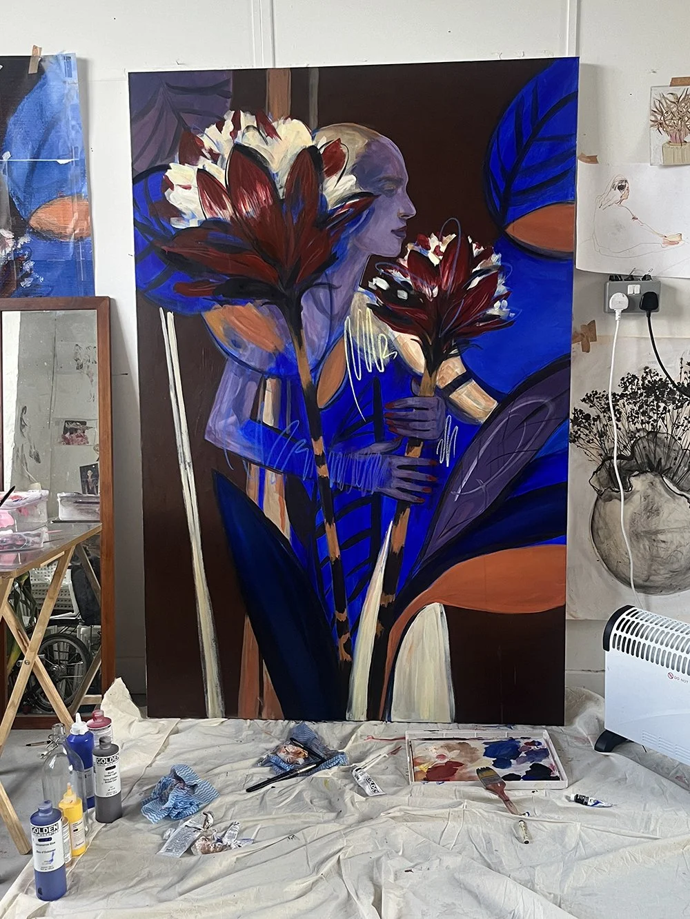 A large mixed media artwork featuring a woman with light skin and long hair, surrounded by bold, colorful flowers and abstract shapes in blue, purple, orange, and red hues, in an artist's studio.