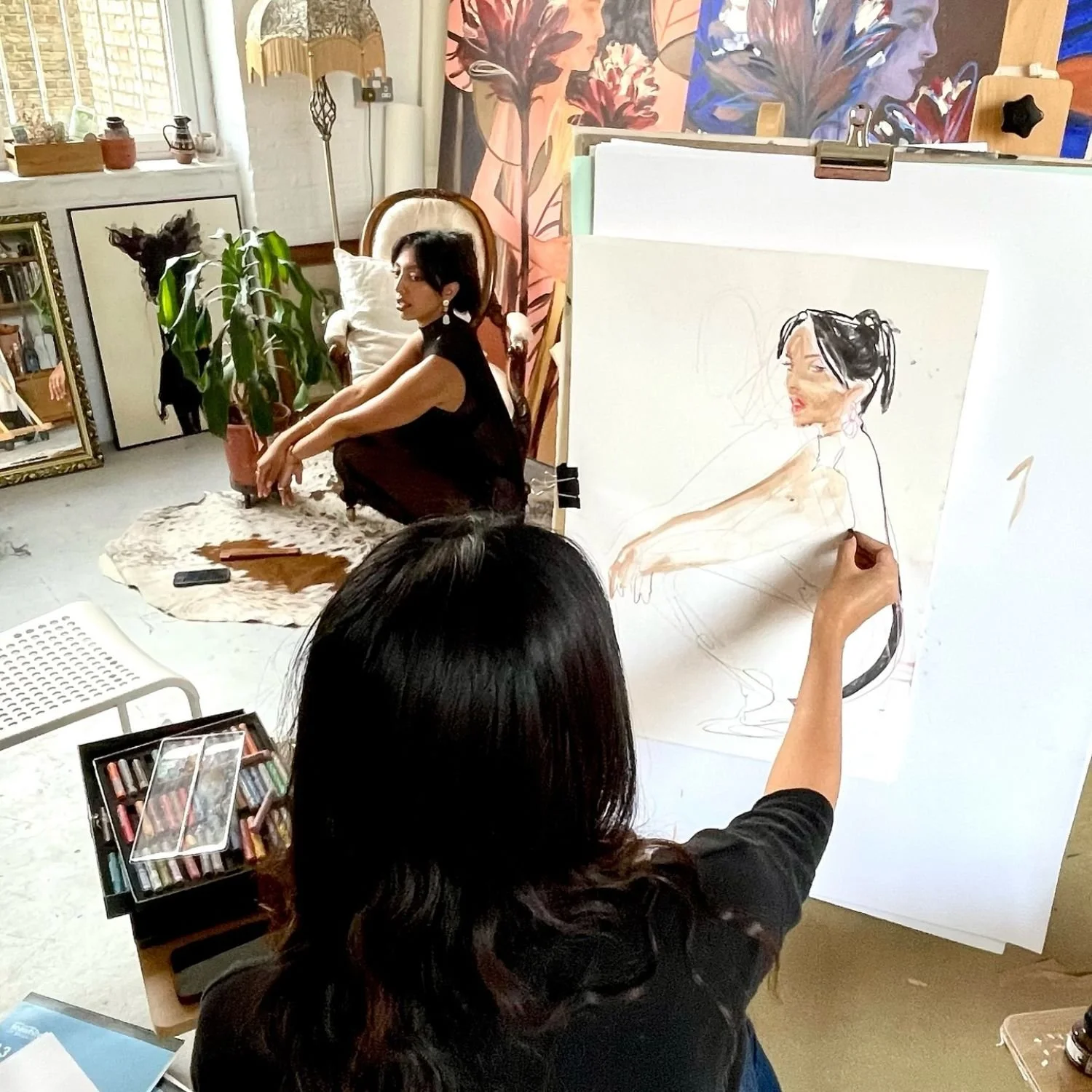 An artist draws a woman sitting on the floor in a studio, working on a portrait of her. The studio has art supplies, paintings, and plants, with a shelf, mirror, and large, colorful paintings in the background.