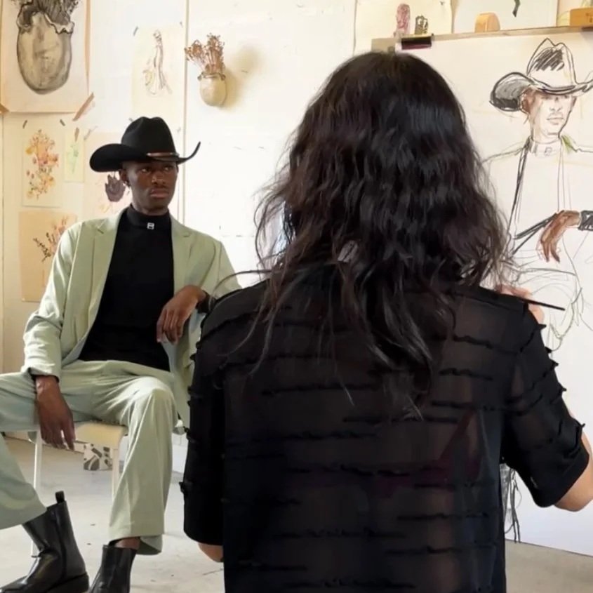 An artist with long dark wavy hair drawing a woman in a hat and holding a gun on a large sketchbook, with a man in a cowboy hat and light-colored suit sitting and observing in the background surrounded by art supplies and sketches.