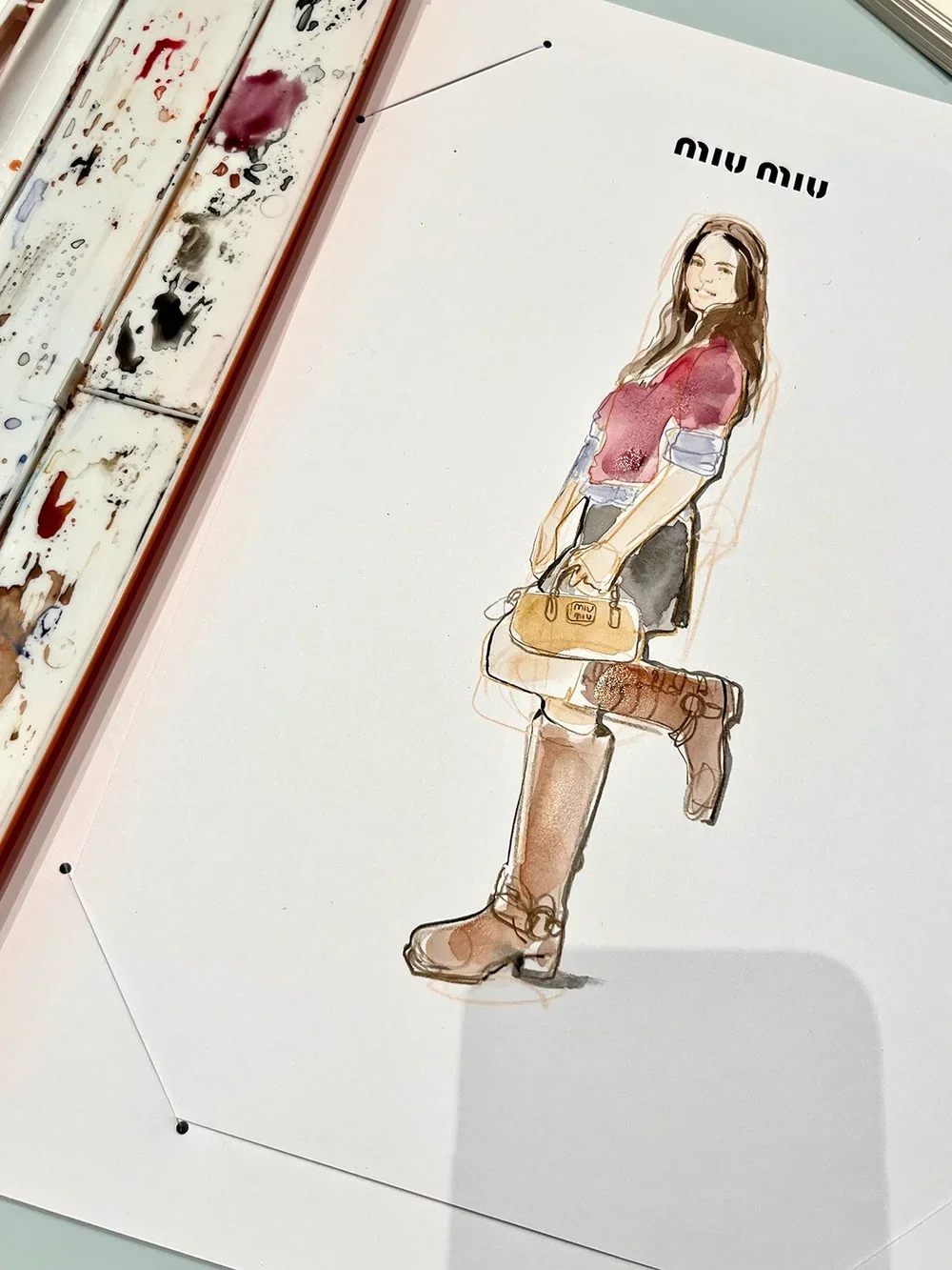 Watercolor fashion illustration of a woman in a red blouse, black skirt, knee-high brown boots, holding a tan purse, from Miu Miu, on white paper with watercolor paints nearby.