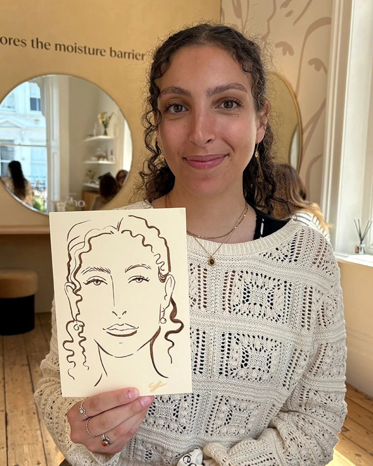 A woman with curly hair and wearing a white crochet sweater is holding a sketch of her face, which shows her with wavy hair, arched eyebrows, and a slight smile.