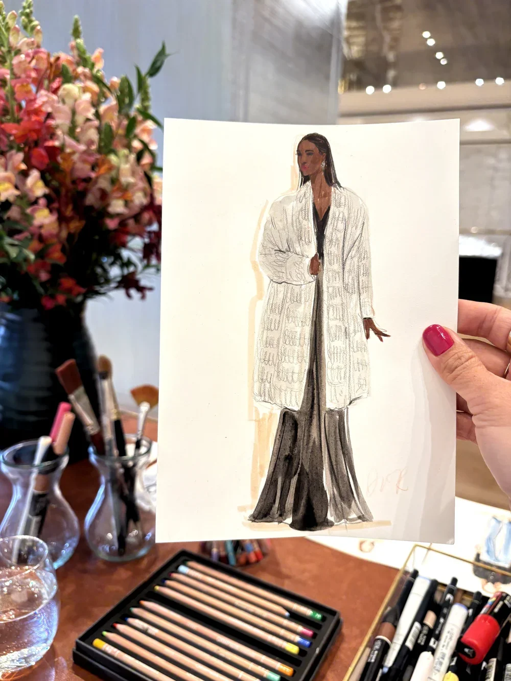 A hand holding a fashion illustration of a woman wearing a long, fur coat and black pants, with a flower arrangement and art supplies on a table in the background.