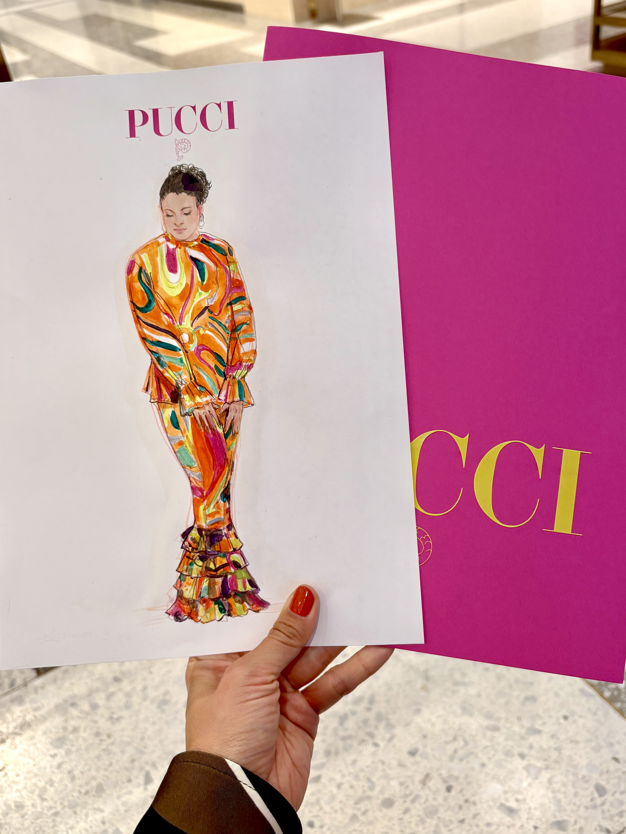 Fashion illustration of a woman in a colorful, abstract patterned outfit with the word "PUCCI" on top, held in a person's hand in front of a magenta display board with "GUCCI" text.