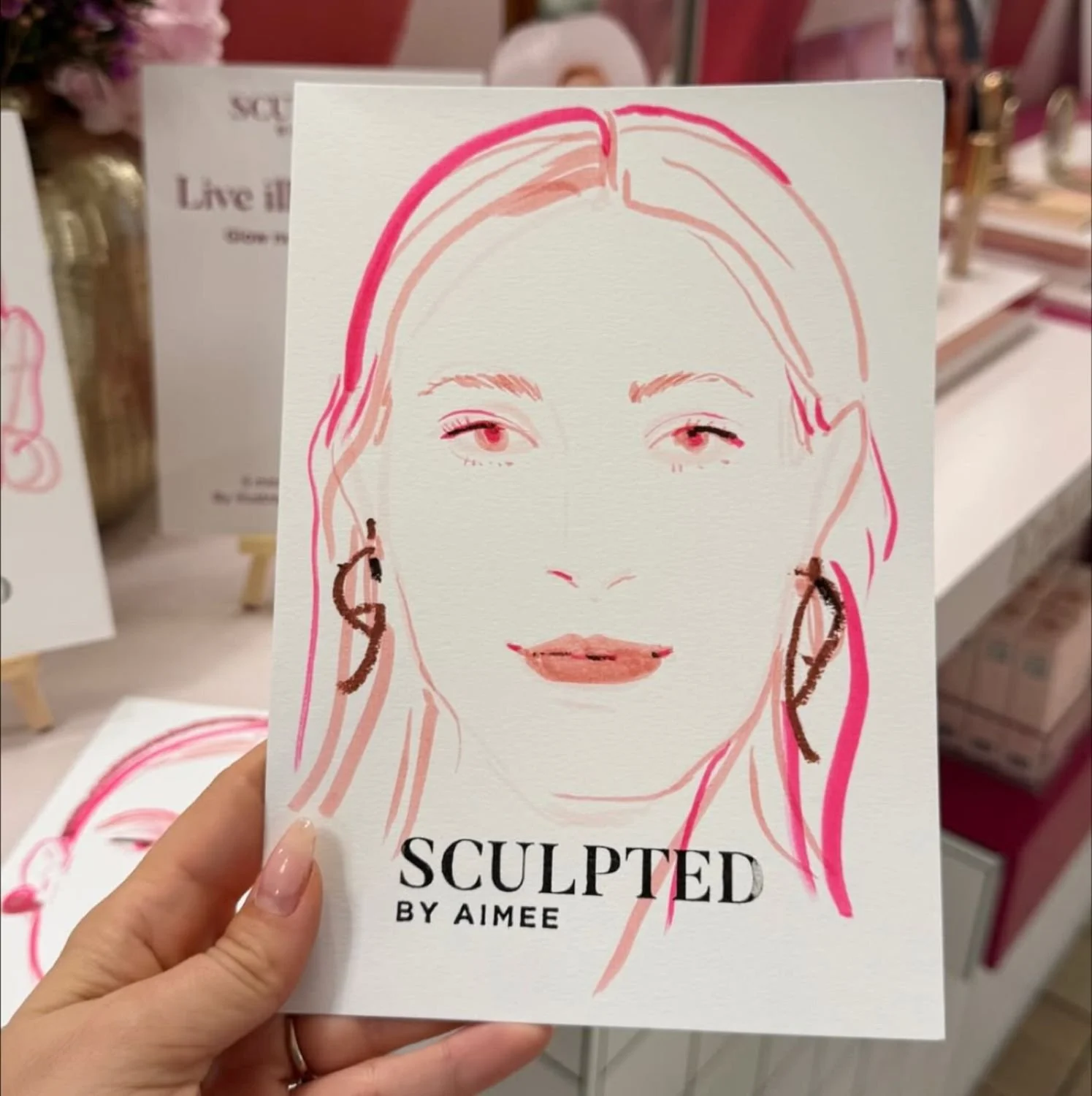 A hand holding a booklet titled 'Sculpted by Aimee' featuring a minimalist line drawing of a woman's face with pink and brown accents.