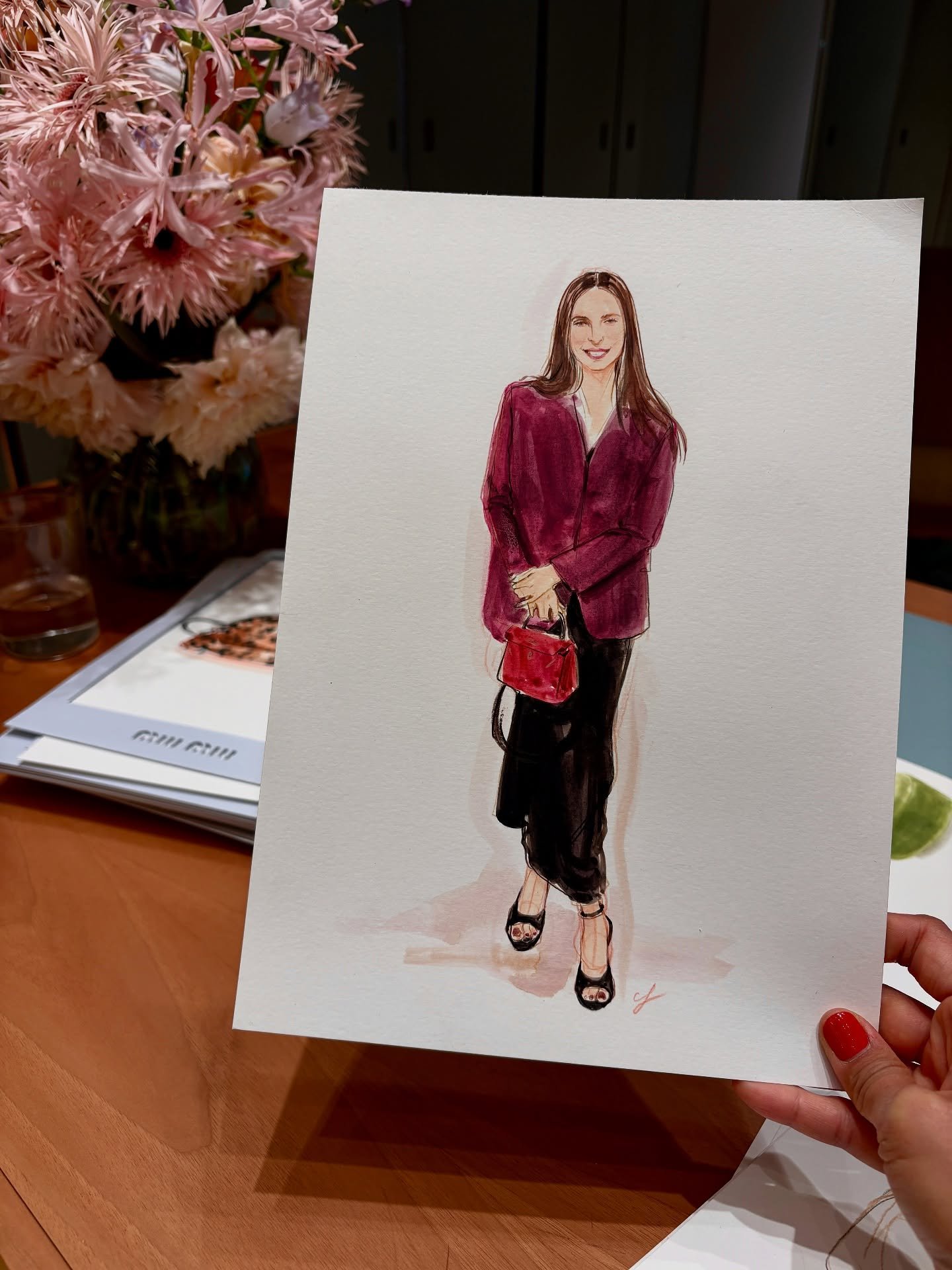 A watercolor illustration of a smiling woman with long brown hair, dressed in a maroon blazer, black pants, and black heels, holding a red handbag.
