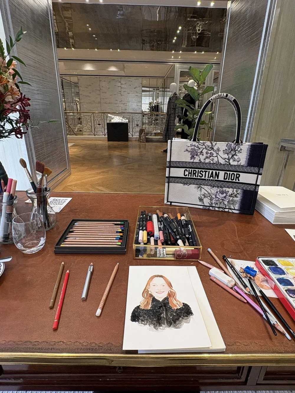 A makeup and art station with watercolor paints, brushes, colored pencils, and a watercolor portrait of a smiling woman on a table. There is a Christian Dior tote bag in the background and a lobby or display area visible through a glass window.