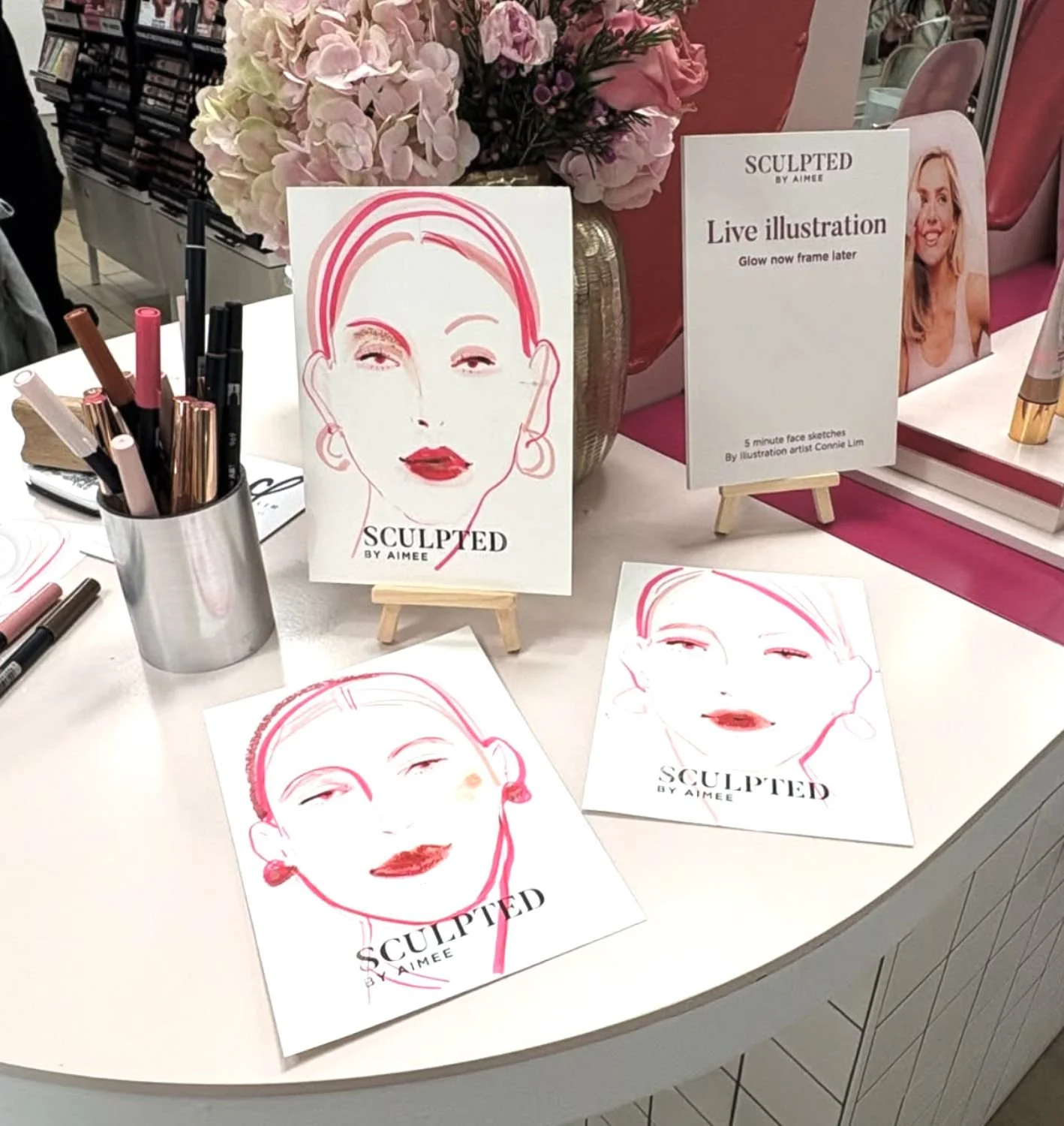 Display of illustrated portrait sketches of a woman's face, with makeup details, on a table at an art event, accompanied by a sign indicating live illustration by Connie Lim.