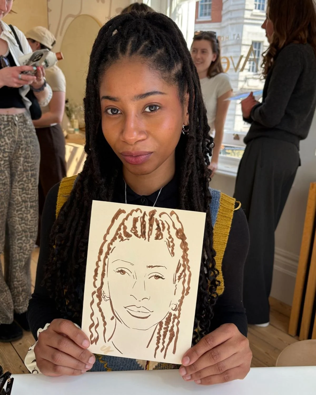 Young woman with long dreadlocks holding a drawing of a woman with dreadlocks, sitting at a table in a well-lit room with large windows.