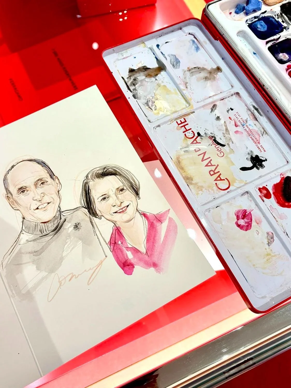 A watercolor painting of a smiling man and woman, with the woman dressed in a pink top, next to an artist's palettes and paint supplies.