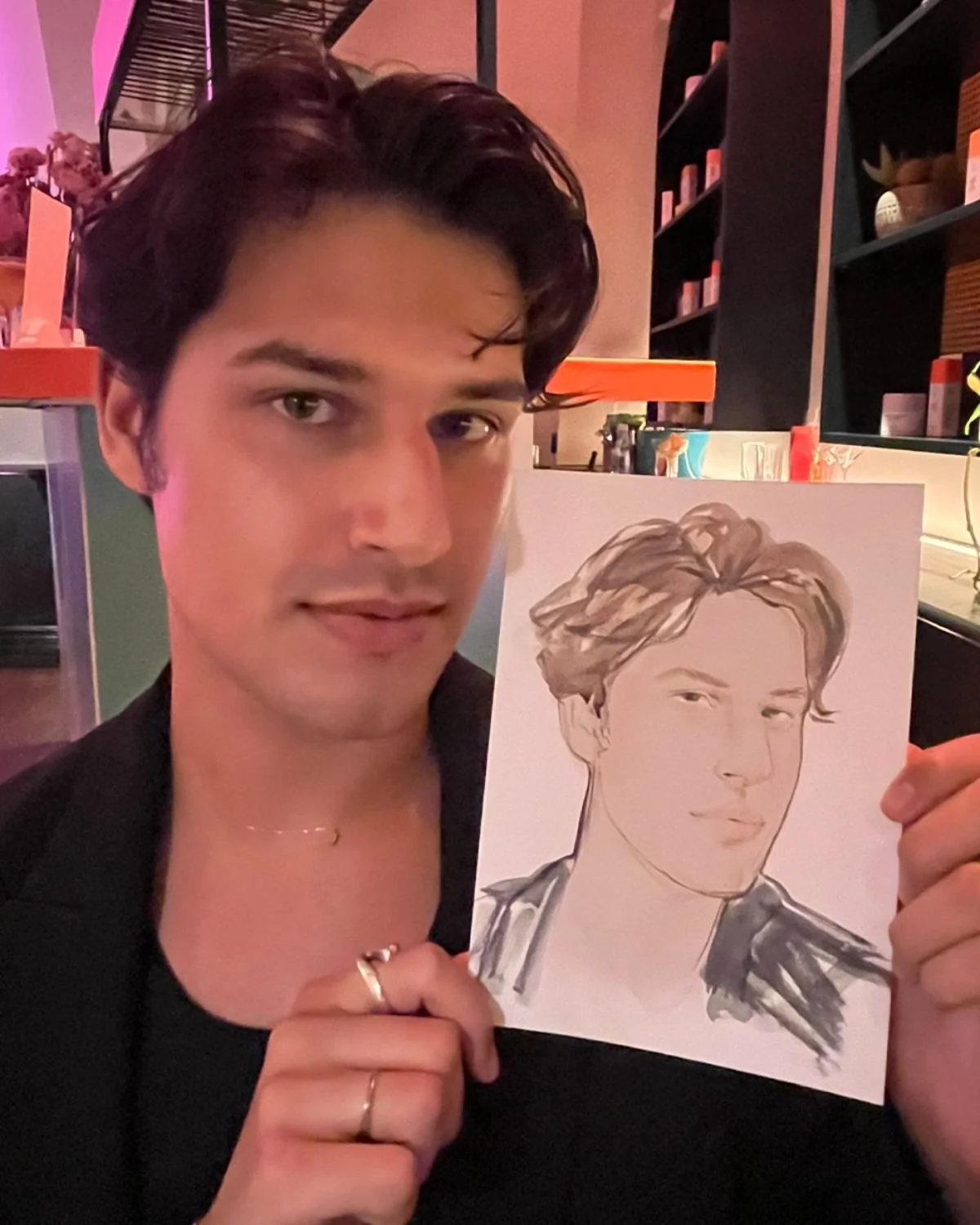 A young man with dark, wavy hair holding a sketch of himself who has similar hair and features, in an indoor setting with shelves and decorative items in the background.