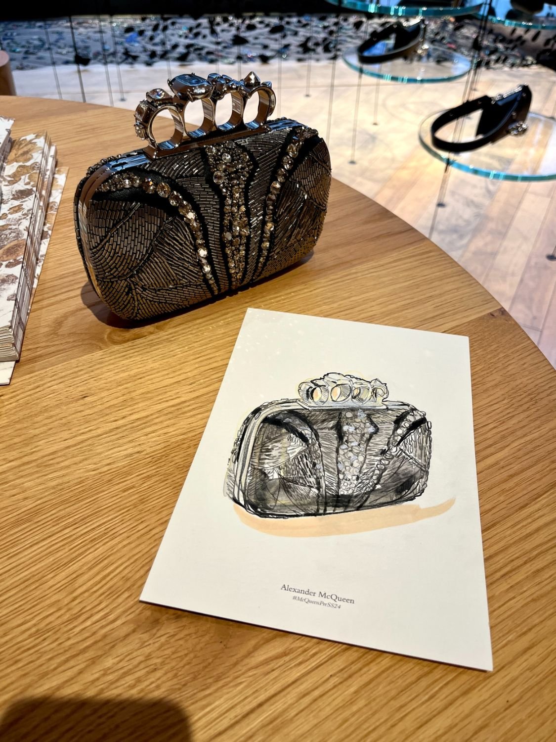 A black and silver jeweled clutch purse with a knuckle ring handle, placed on a round wooden table next to a sketch or card featuring a similar purse illustration. In the background, there are display cases with sunglasses and other accessories.
