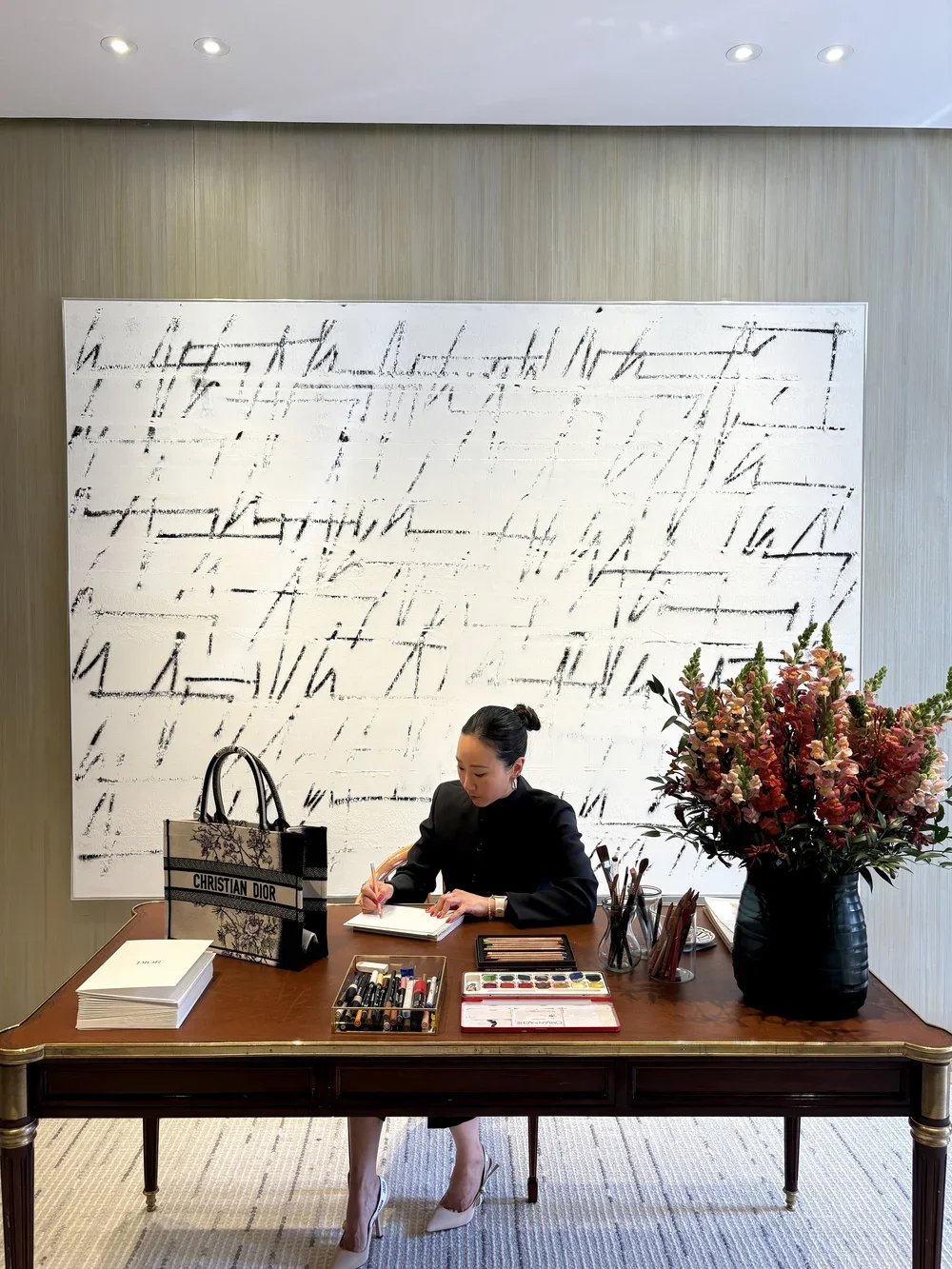 A woman sitting at a wooden desk with art supplies, a Christian Dior shopping bag, and large flower arrangement, writing in a notebook in a modern, well-lit room with abstract artwork on the wall.