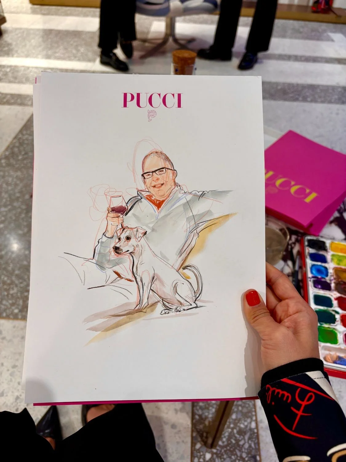 Hand holding a fashion illustration on paper with the word "Pucci" at the top. The illustration depicts a man with glasses smiling, holding a glass of red wine, sitting on a chair with a dog sitting on the ground beside him. Art supplies and a pink b
