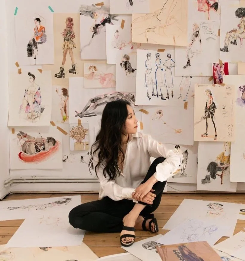 A woman sitting on the floor surrounded by fashion sketches and drawings on the wall and scattered on the floor.