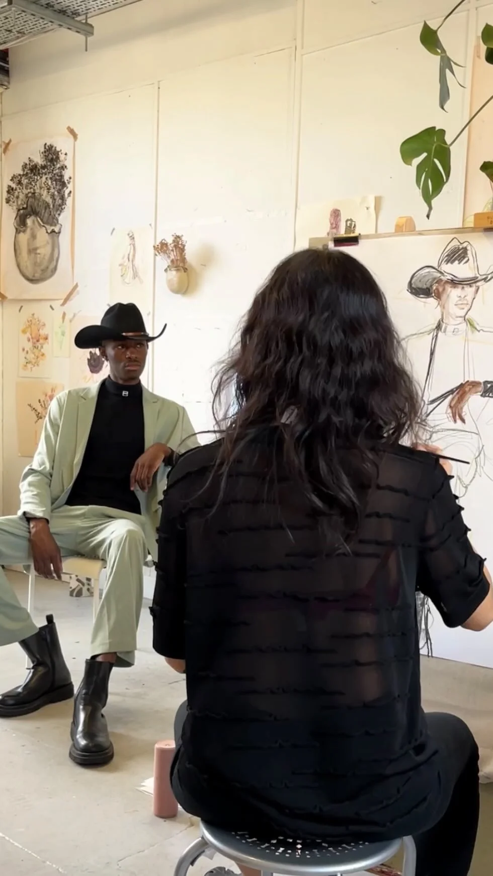 A woman with dark, wavy hair painting a sketch of a cowboy woman wearing a hat in an art studio. A man in a cowboy hat and western attire sitting on a chair, observing. The studio walls are decorated with drawings and paintings, including vases and flowers.