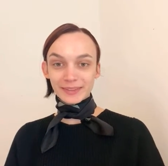 Young woman with short brown hair and light skin, wearing a black sweater and a black scarf around her neck, standing against a plain beige wall, smiling slightly at the camera.