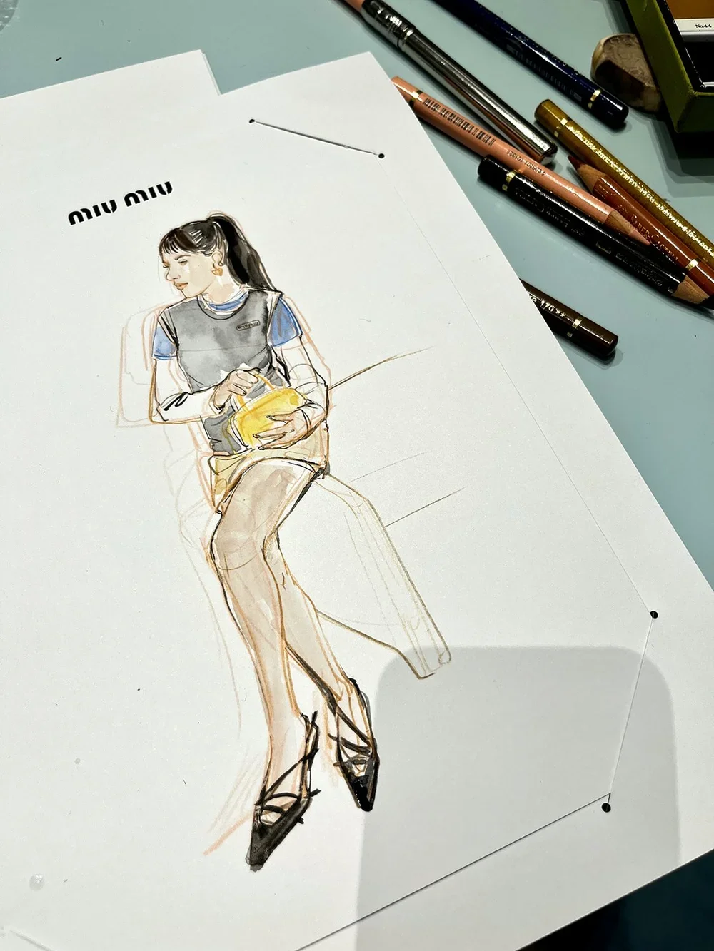 Fashion sketch of a woman sitting on a chair, holding a yellow purse, with art supplies nearby.