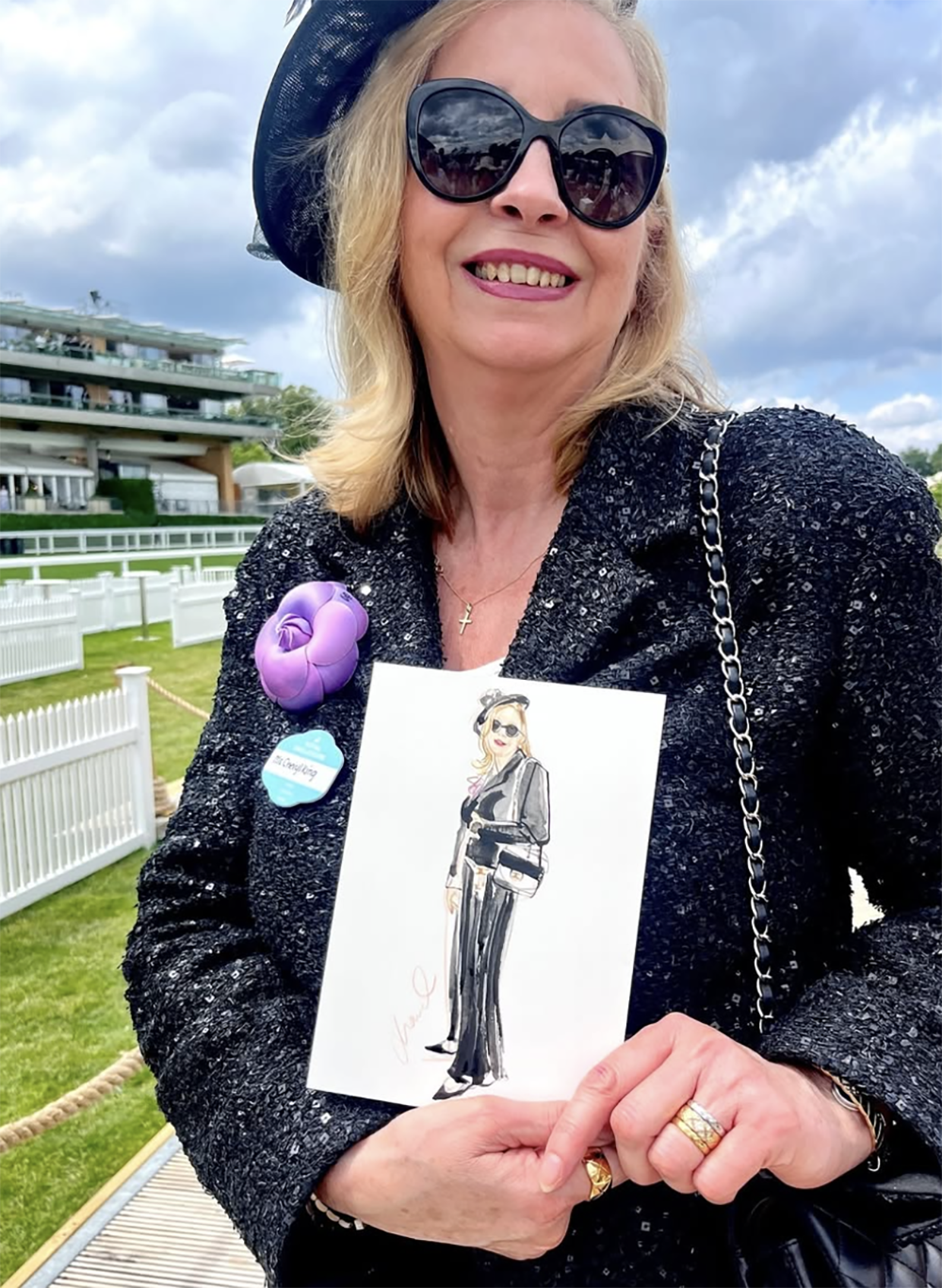 Smiling woman wearing large sunglasses, black hat, black sparkly jacket, and gold jewelry, holding a fashion illustration of her in a black suit, with buttons and a handbag. She stands outdoors near a racetrack or similar venue, with a white fence an