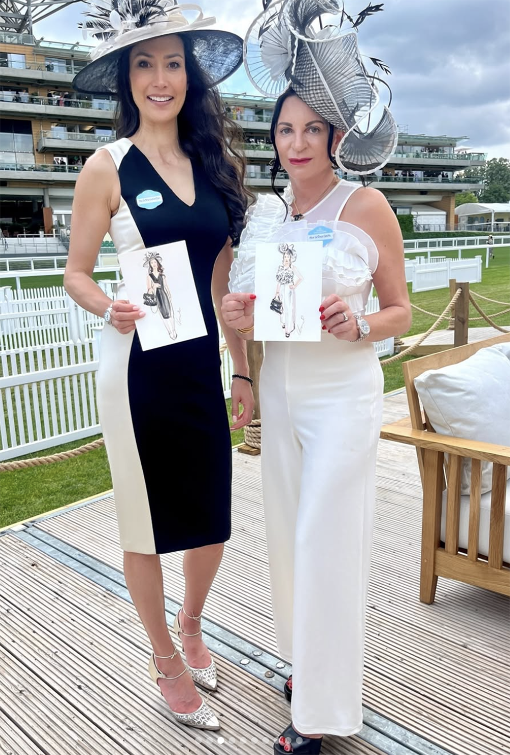 Two women in elegant outfits and large decorative hats, holding fashion sketches, standing outdoors near a wooden fence and lush green lawn with a multi-level building in the background.