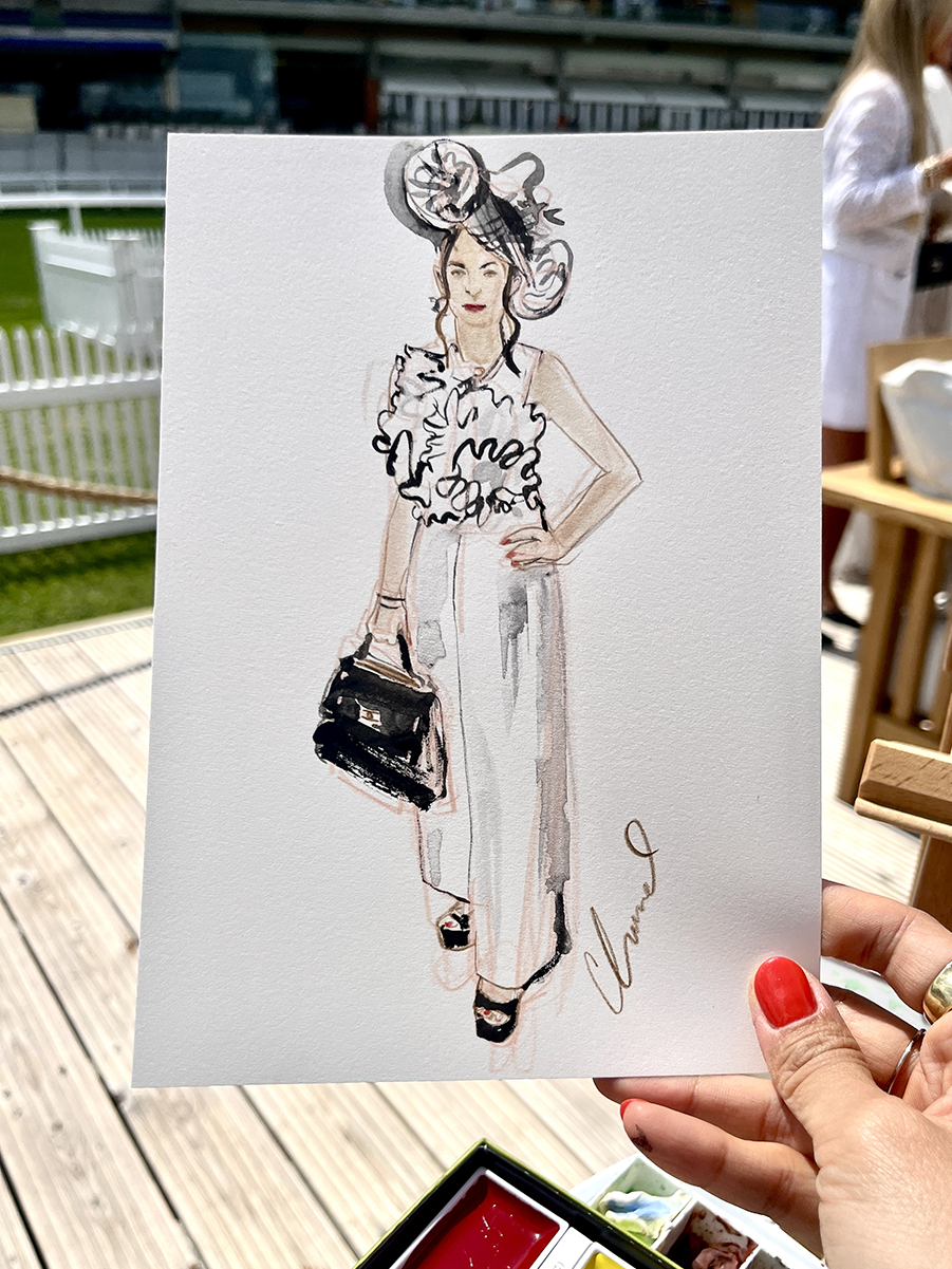 A hand holding a watercolor illustration of a woman with a large hat, ruffled top, wide-leg pants, and a handbag, outdoors on a wooden deck.