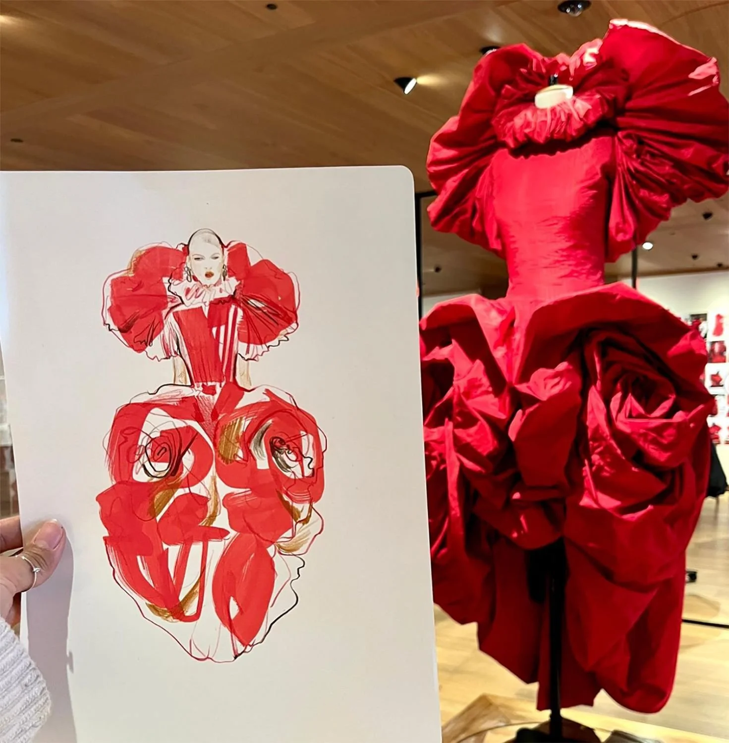 Sketch of a woman wearing an elaborate red dress, held in front of a real red dress on a mannequin display at alexander mcqueen