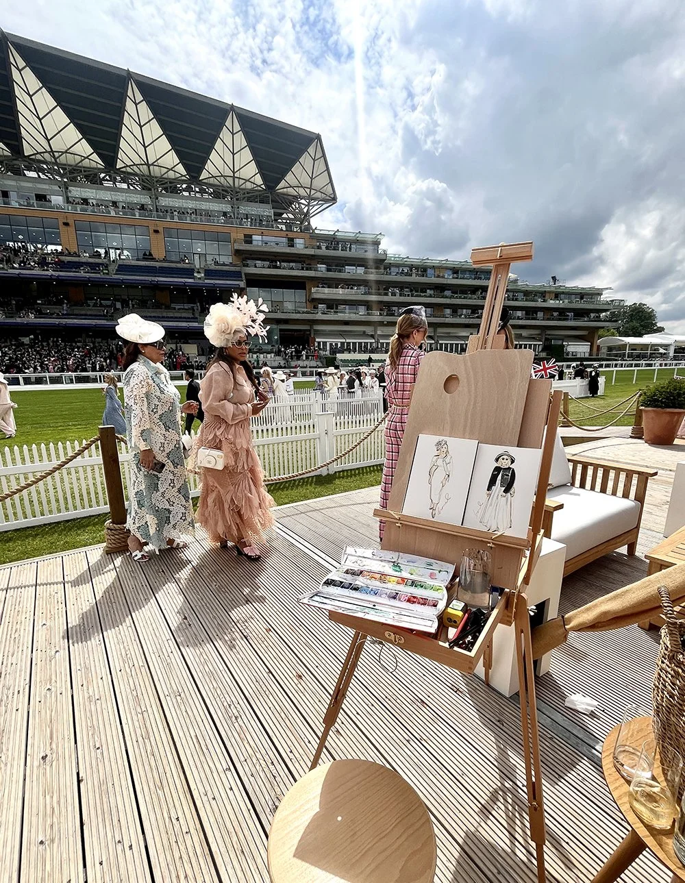 Fashionable women in elegant outfits walking on a wooden deck at a racecourse, with a modern building and cloudy sky in the background. An artist's easel with sketches and painting supplies is in the foreground.