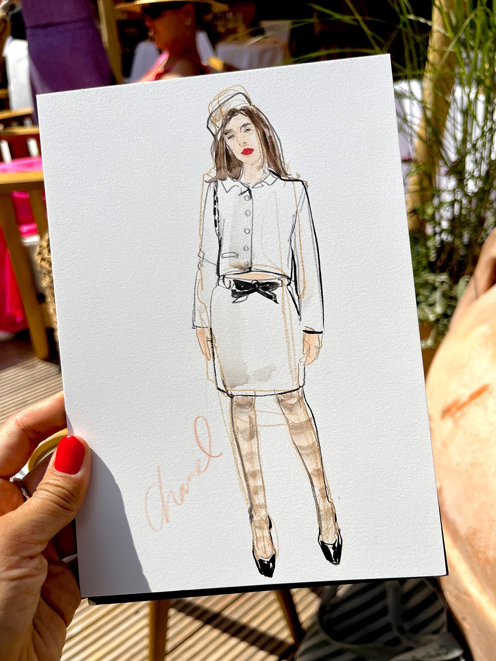 Illustration of a woman wearing a white blouse, a white skirt with a black belt, plaid stockings, and black heels. The woman has brown hair, red lipstick, and is wearing a hat. The illustration is on a white canvas, held by a person with red nail pol