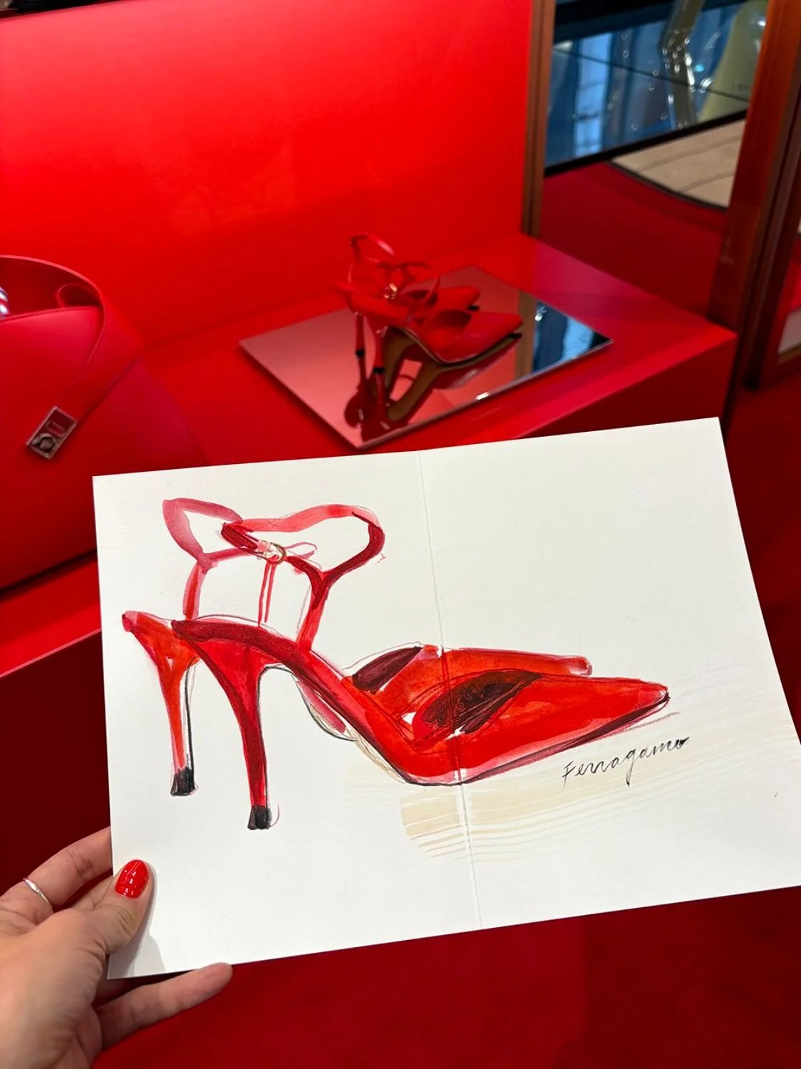 A colorful illustration of a red high-heeled sandal with ankle straps. The illustration is being held in front of a display of the same red sandal in a ferragamo store.