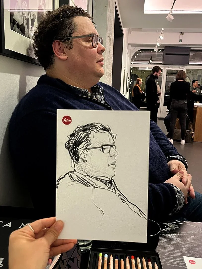 A person sitting at a cafe holding a sketchbook with a portrait sketch of a man with glasses and curly hair. The sketch appears in the style of a line drawing. The person with the sketchbook is not the same as the man in the photograph and they are i