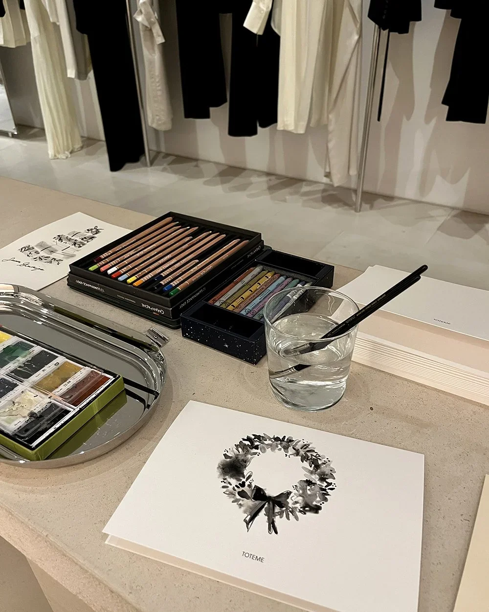 A workspace with colored pencils, watercolor paints, a glass of water with a paintbrush, and a greeting card with a monochrome floral wreath design for Toteme in-store activation.