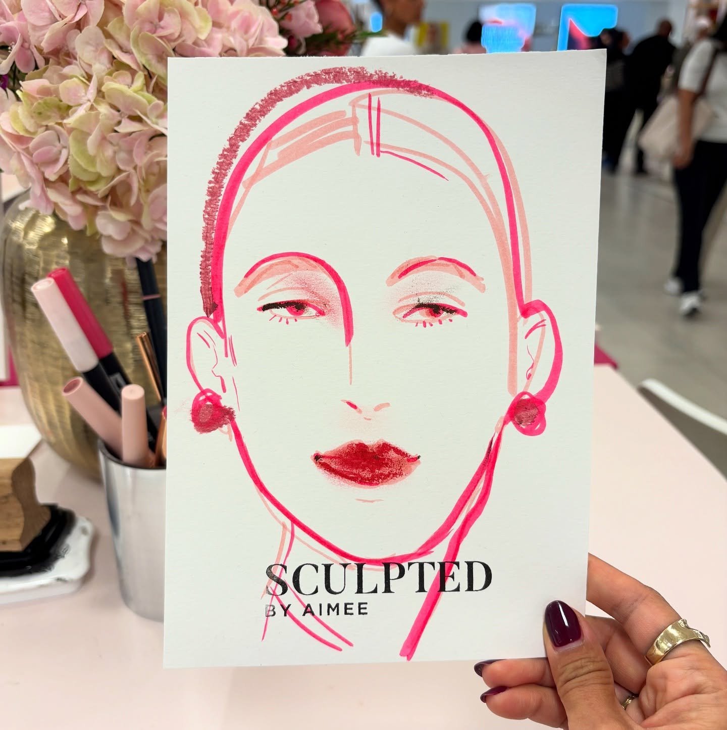 A promotional card with a stylized line drawing of a woman's face in pink and red. The drawing features a woman with earrings, lipstick, and half-closed eyes. The card has the text "SCULPTED BY AIMEE" at the bottom. The background includes a pink flower arrangement and blurred people in a busy indoor setting.
