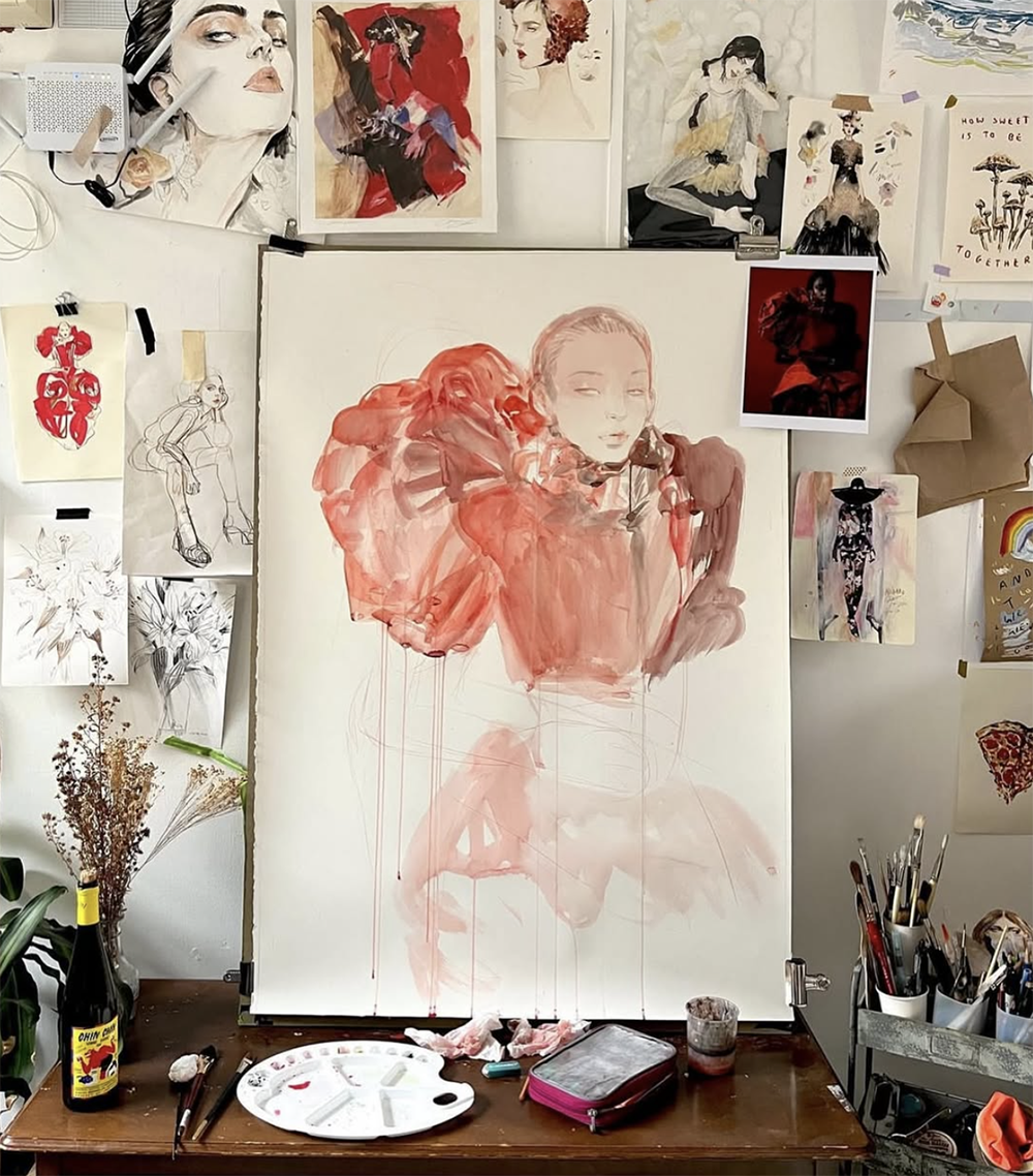 A work-in-progress watercolor painting of a woman with fiery red hair and a large, flowing red dress, on a white easel in an art studio surrounded by sketches, photos, and art supplies.