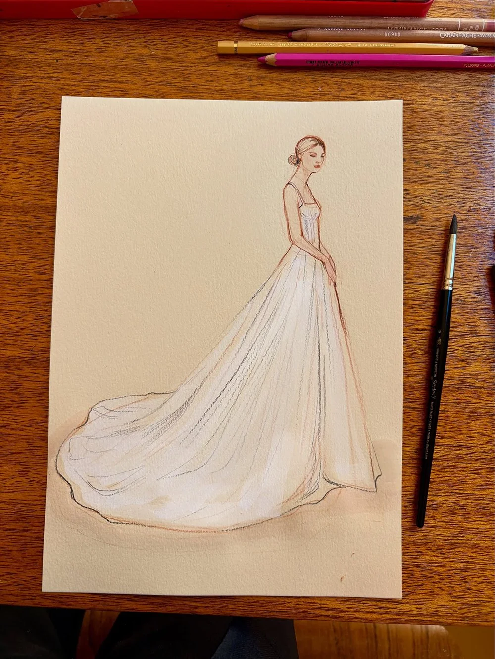 A sketch of a woman wearing a long wedding dress, drawn on textured paper with colored pencils and a pen nearby, on a wooden table.
