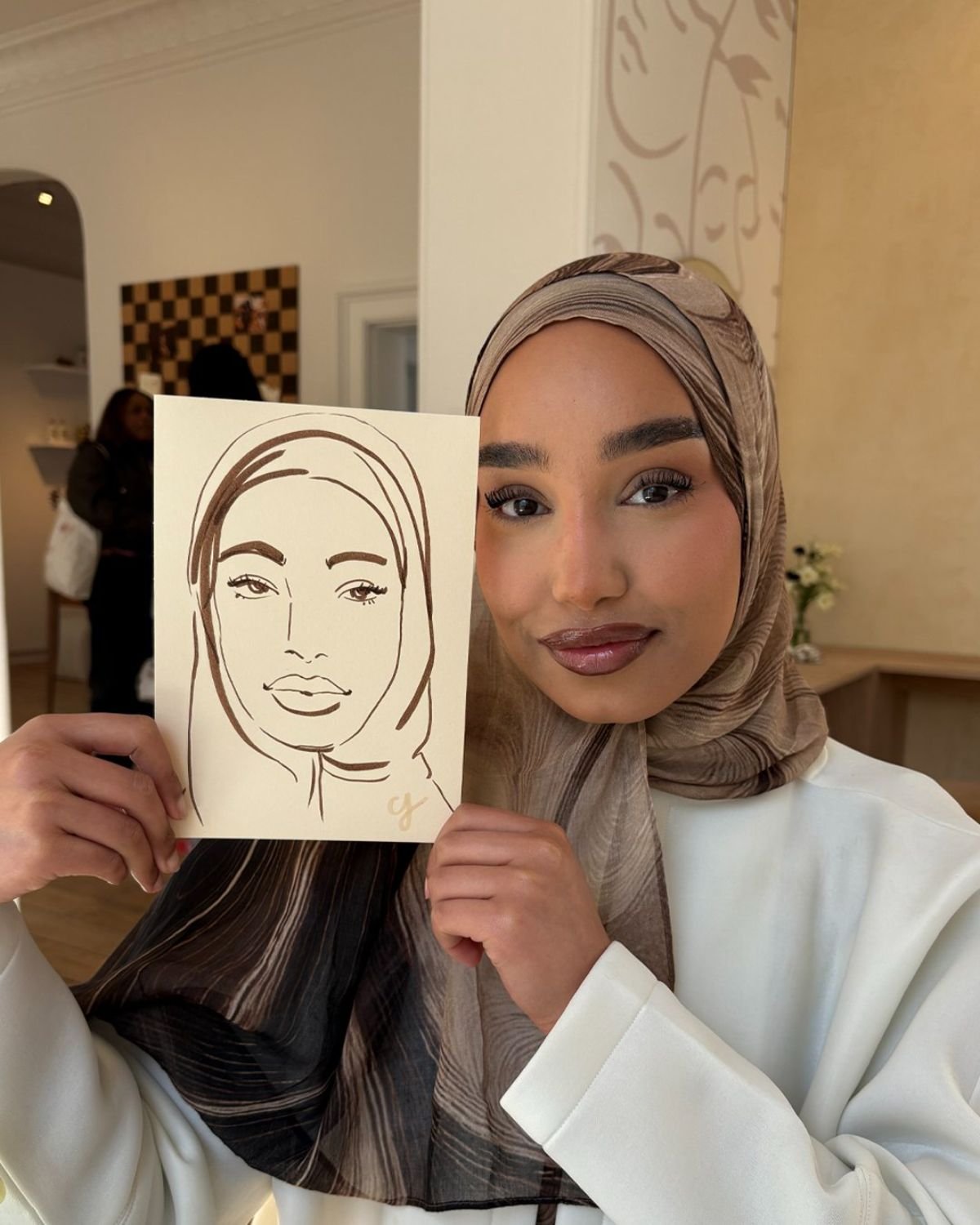 A woman in a beige headscarf and white blazer holding a drawing of herself in front of her face, smiling at the camera.