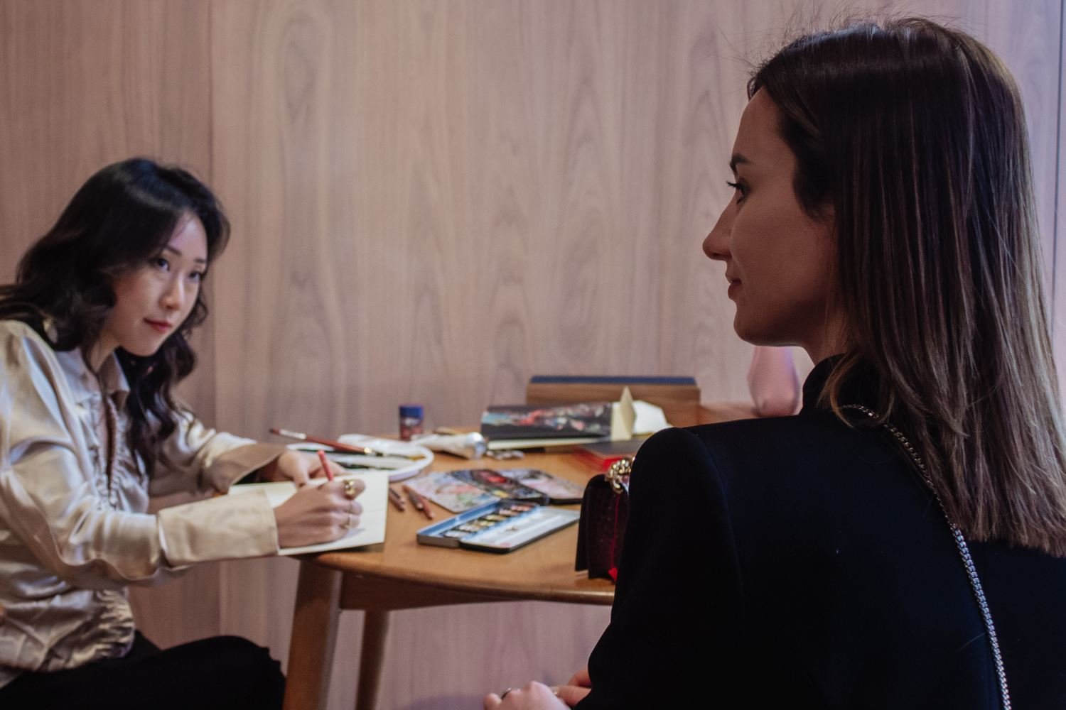Two women sitting at a wooden table with art supplies, engaged in conversation, in a room with wood-paneled walls.