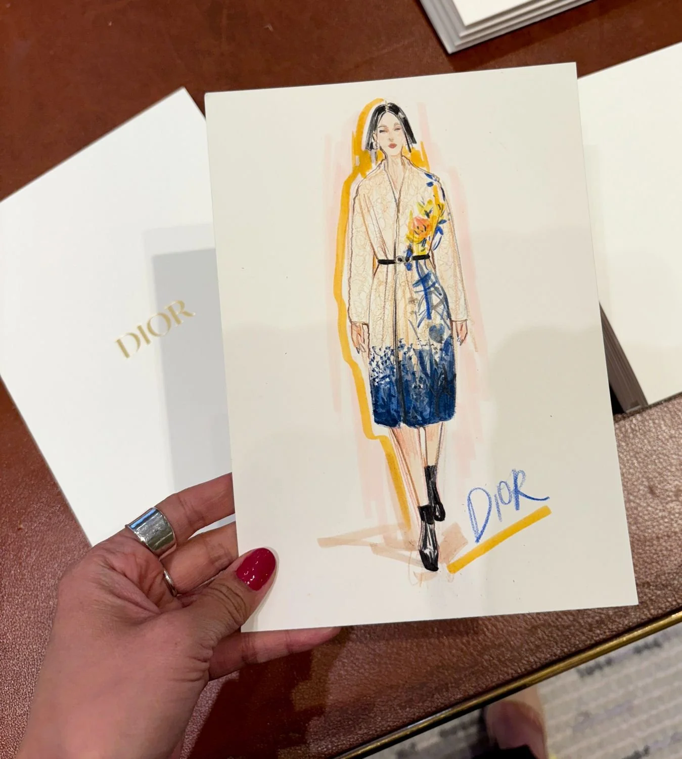 Fashion illustration of a woman wearing a floral patterned coat with a beige and blue gradient, black belt, and black ankle boots, with the Dior logo written in blue and yellow below.
