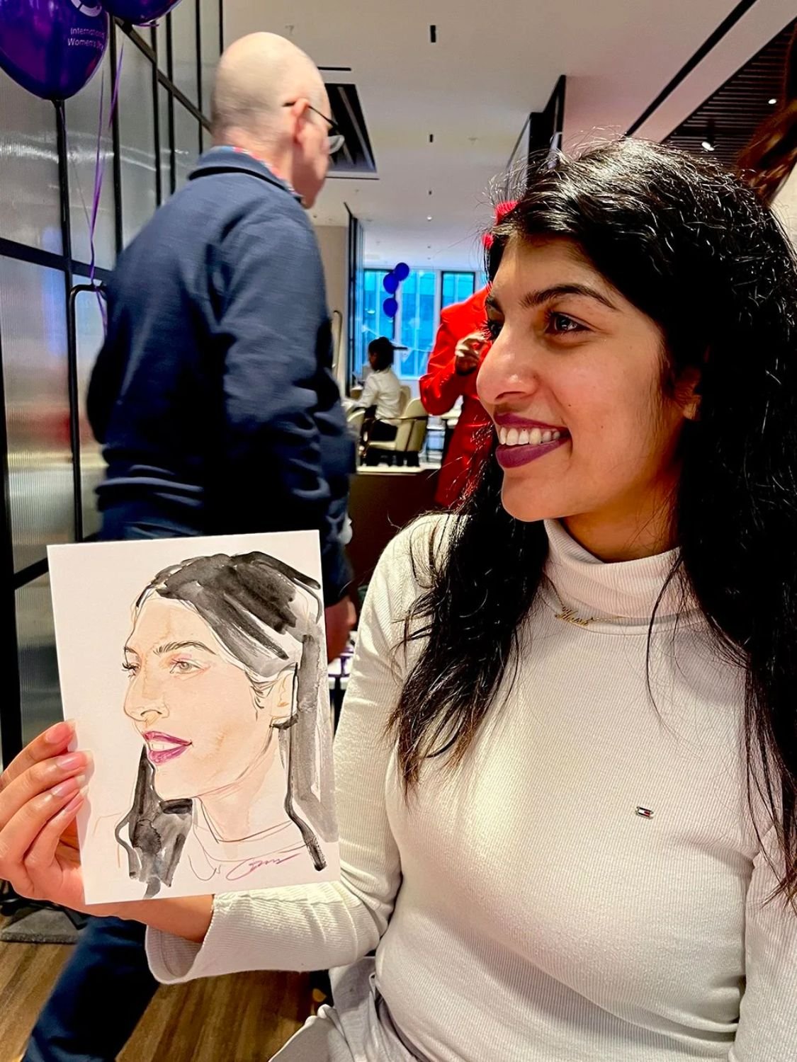 A woman with dark curly hair smiling, holding a portrait sketch of herself at an indoor event.