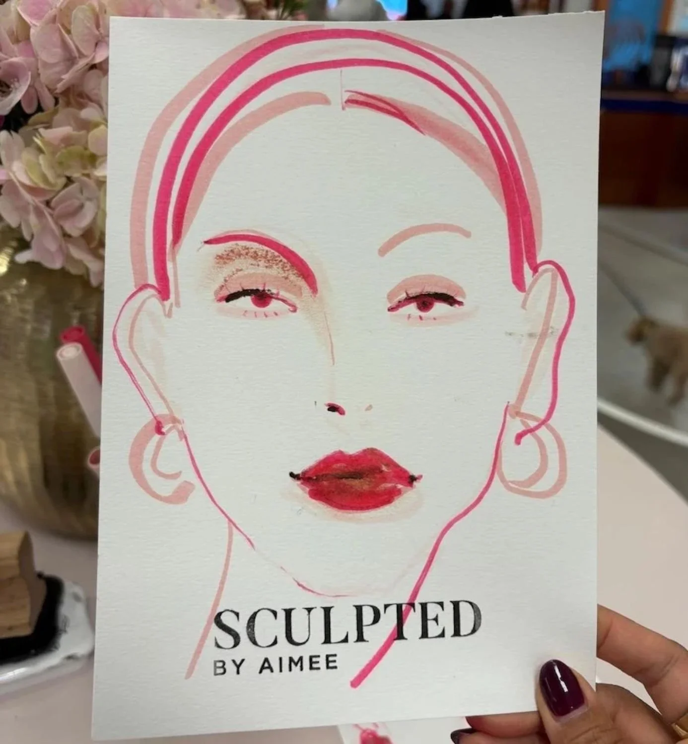 A sketch of a woman's face with pink lines outlining her hair and facial features, with artful use of lipstick and eye makeup. The drawing is part of a promotional card titled 'Sculpted by Aimee'.