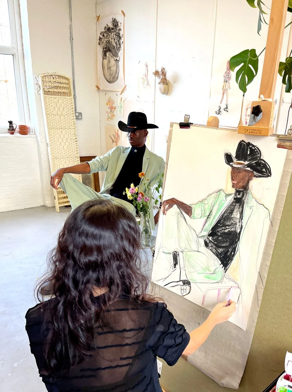 An artist creating a drawing of a person seated in a chair, wearing a black hat and glasses, with artwork and plants in the background.