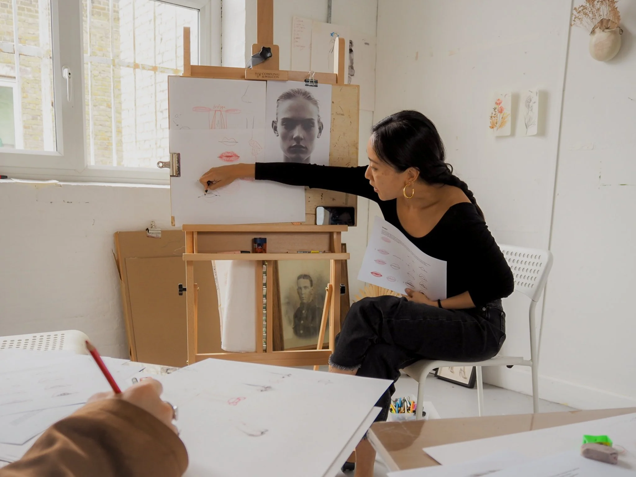 A woman is sitting on a chair while pointing at a drawing of a face on a whiteboard, which includes sketches of lips and a nose. She is holding a piece of paper with similar sketches. The background features artwork and a window.