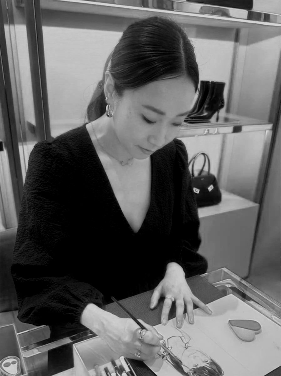 A woman with dark hair styled back, wearing earrings, a necklace, and a dark top, is sitting at a table drawing a portrait of a woman on paper with colored pencils or pens. A heart-shaped object is on the table, and a train of shelves and a bag are in the background.