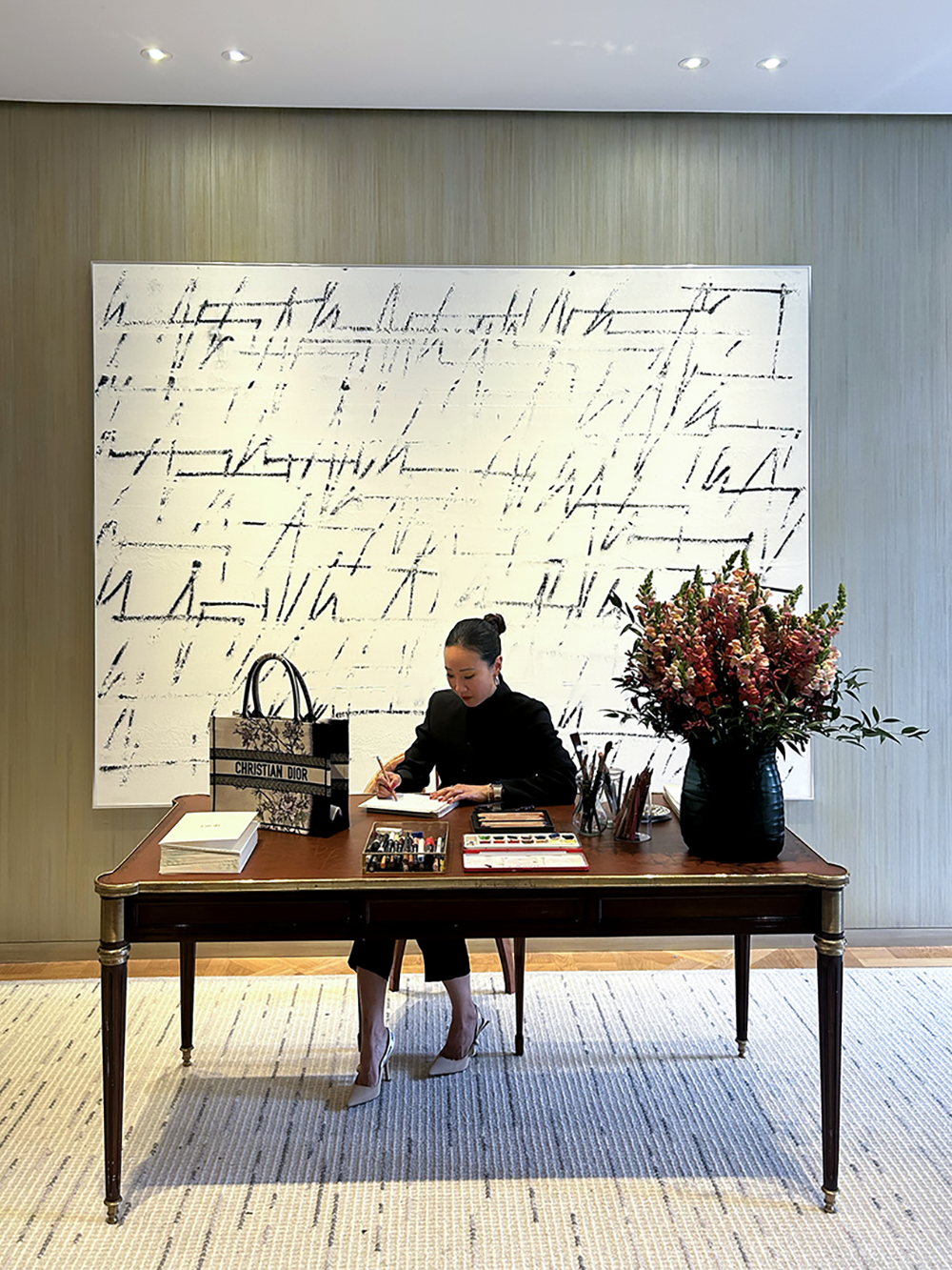 A woman in black attire sitting at a wooden desk, writing in a notebook, with a Christian Dior bag, a large bouquet of pink flowers, and art supplies on the desk, in a modern interior space.