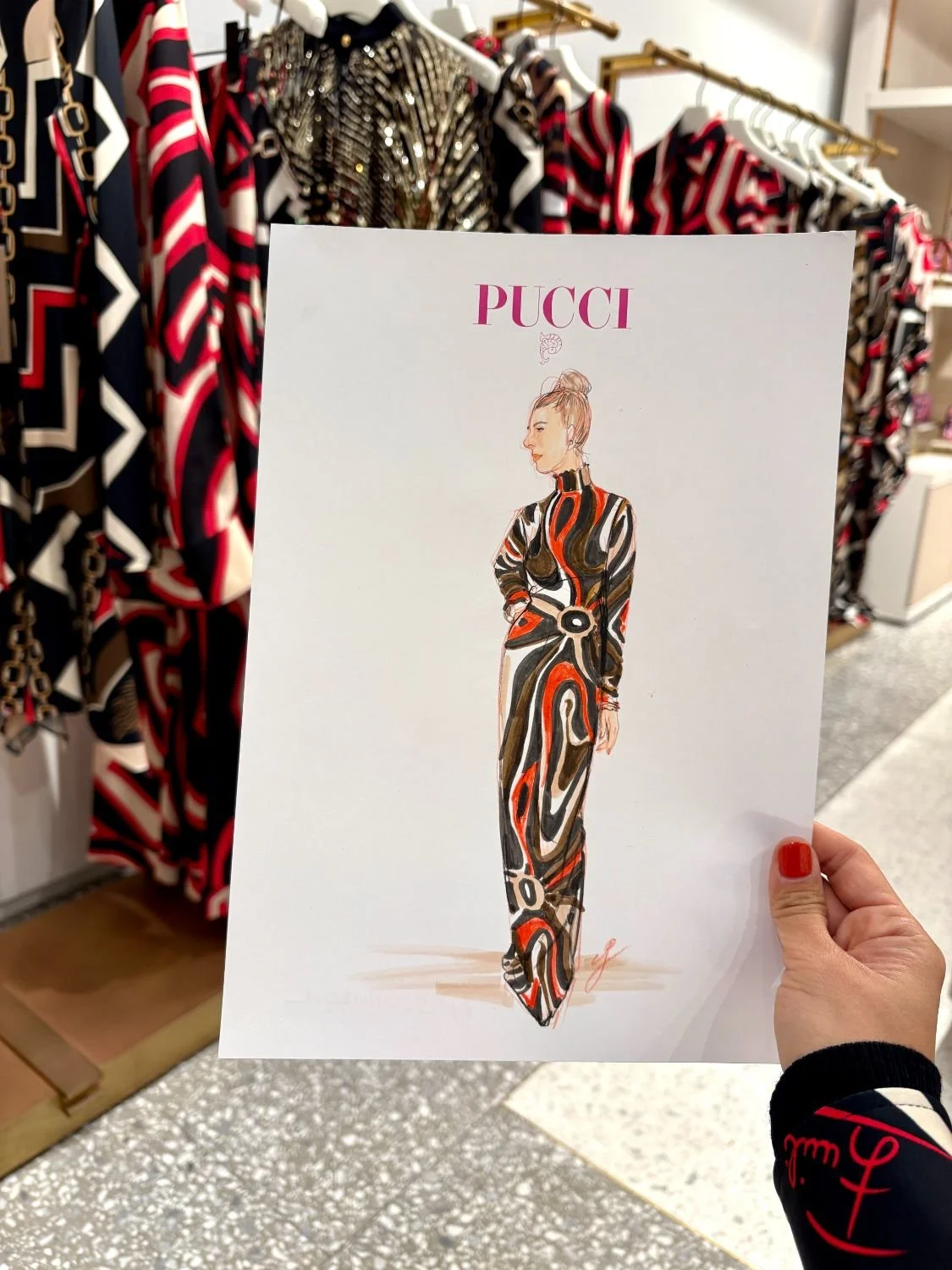 A person holding a fashion illustration of a woman wearing a long, patterned dress in front of a clothing rack with similar patterned garments.