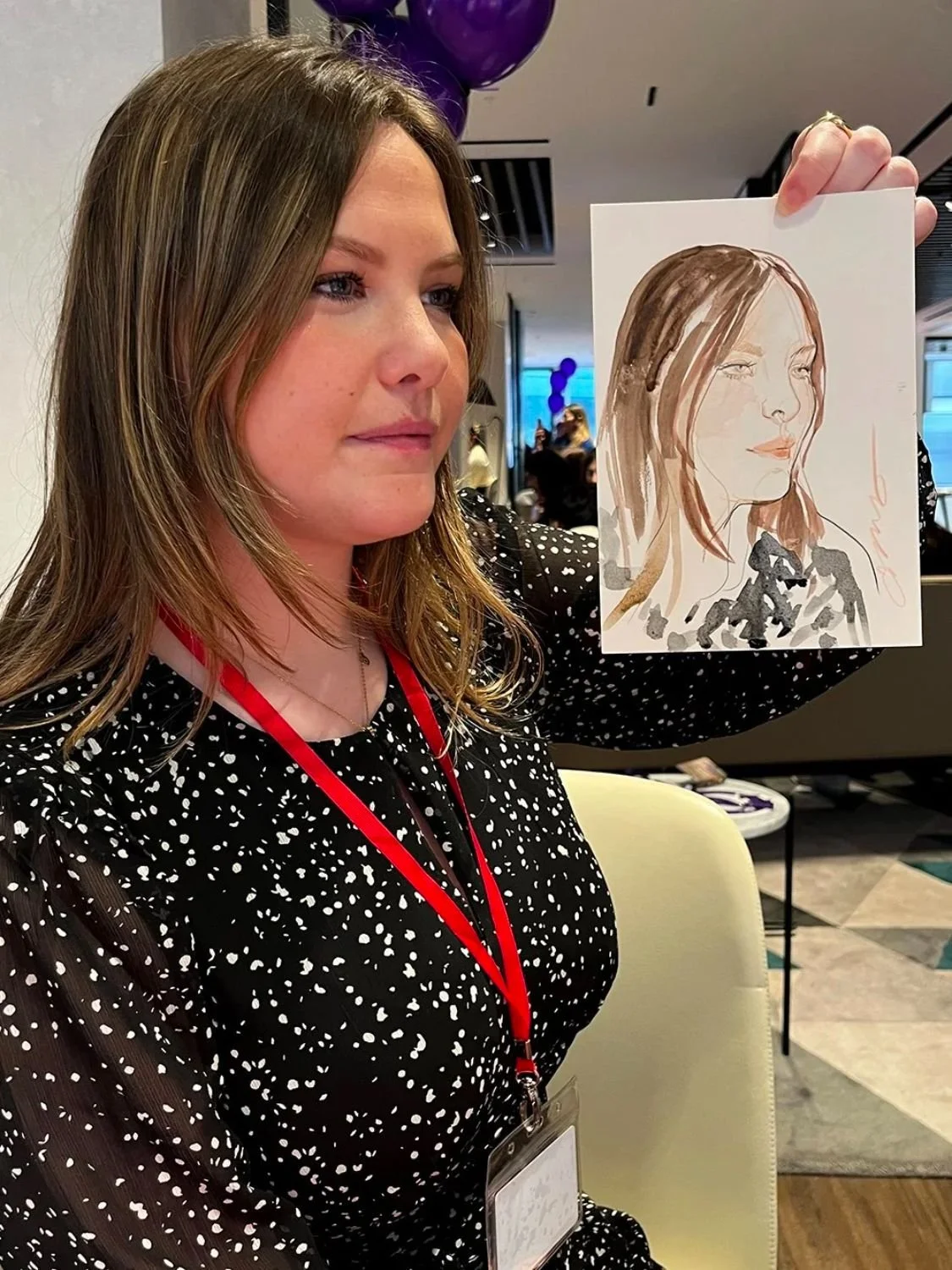 A woman with light brown hair holds up a watercolor portrait of herself, showing her side profile with her hair parted to the side, dressed in a patterned top, sitting in a room with balloons and other people in the background.